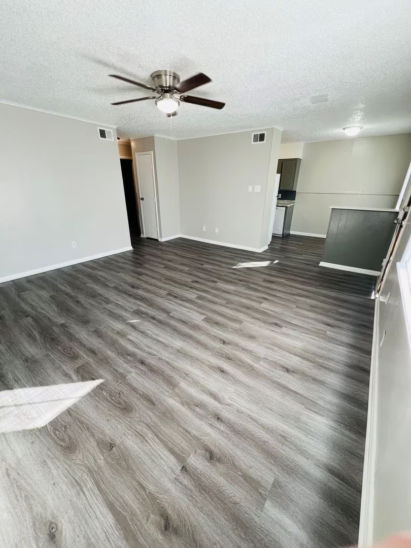 Empty living room with gray wood floors, white walls, ceiling fan, and a doorway to a kitchen.
