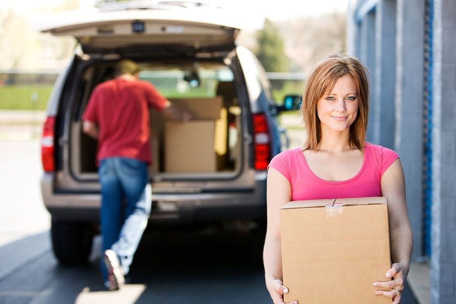 Woman smiling, holding a box, watches as a man loads boxes into a car.