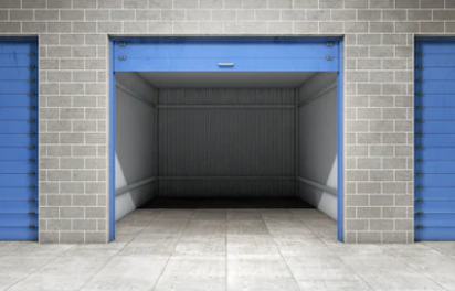 An empty storage unit with a blue door open, flanked by closed blue doors, in a gray brick building.