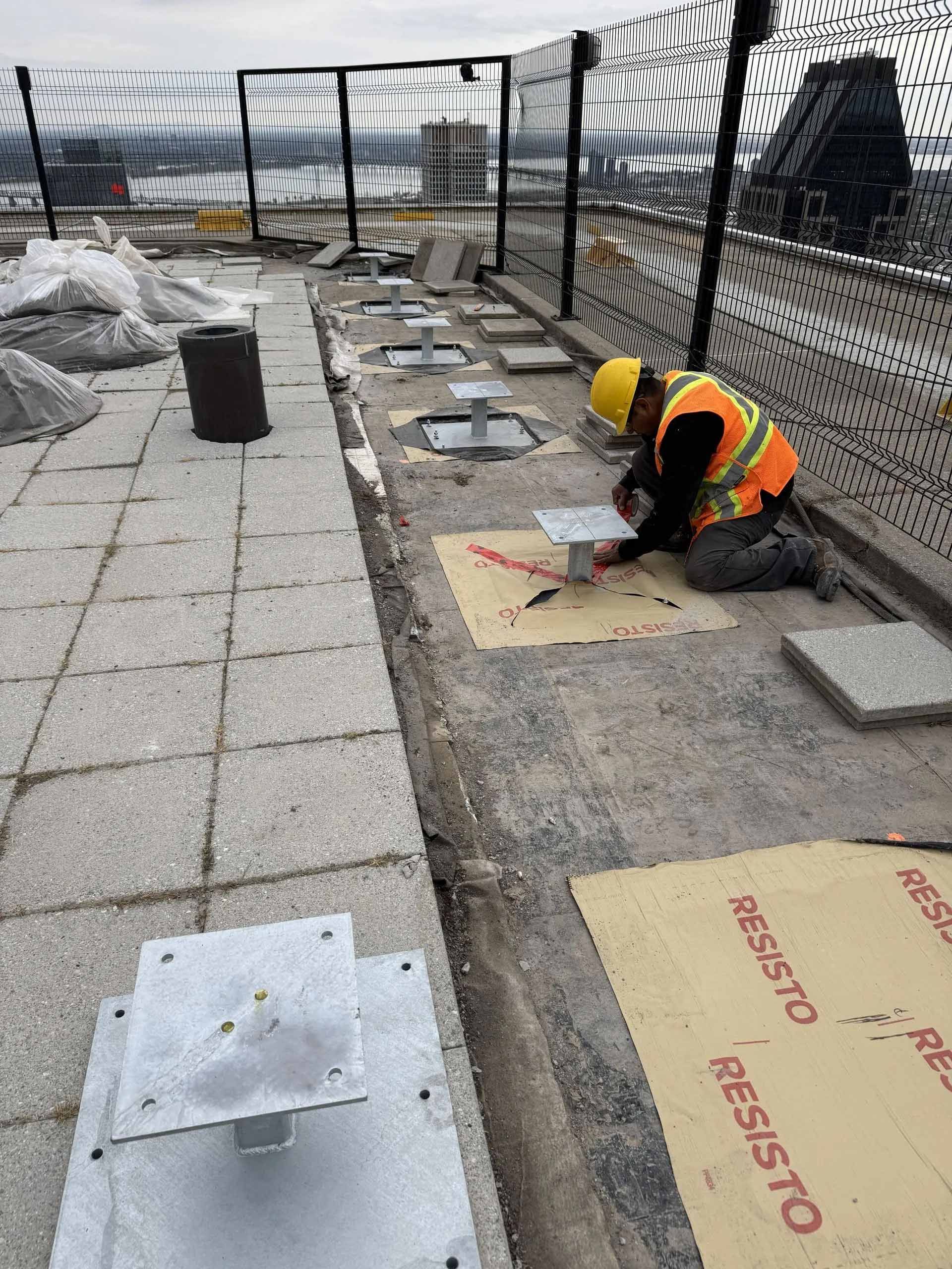 Un ouvrier du bâtiment installe une base métallique sur un toit, portant un gilet de sécurité et un casque de chantier.