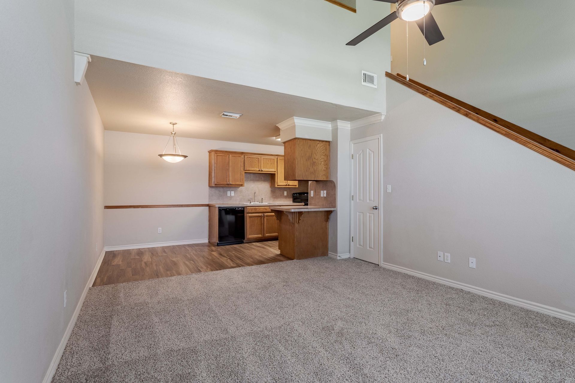 Living room with kitchen in background, neutral colors, carpet, high ceilings.