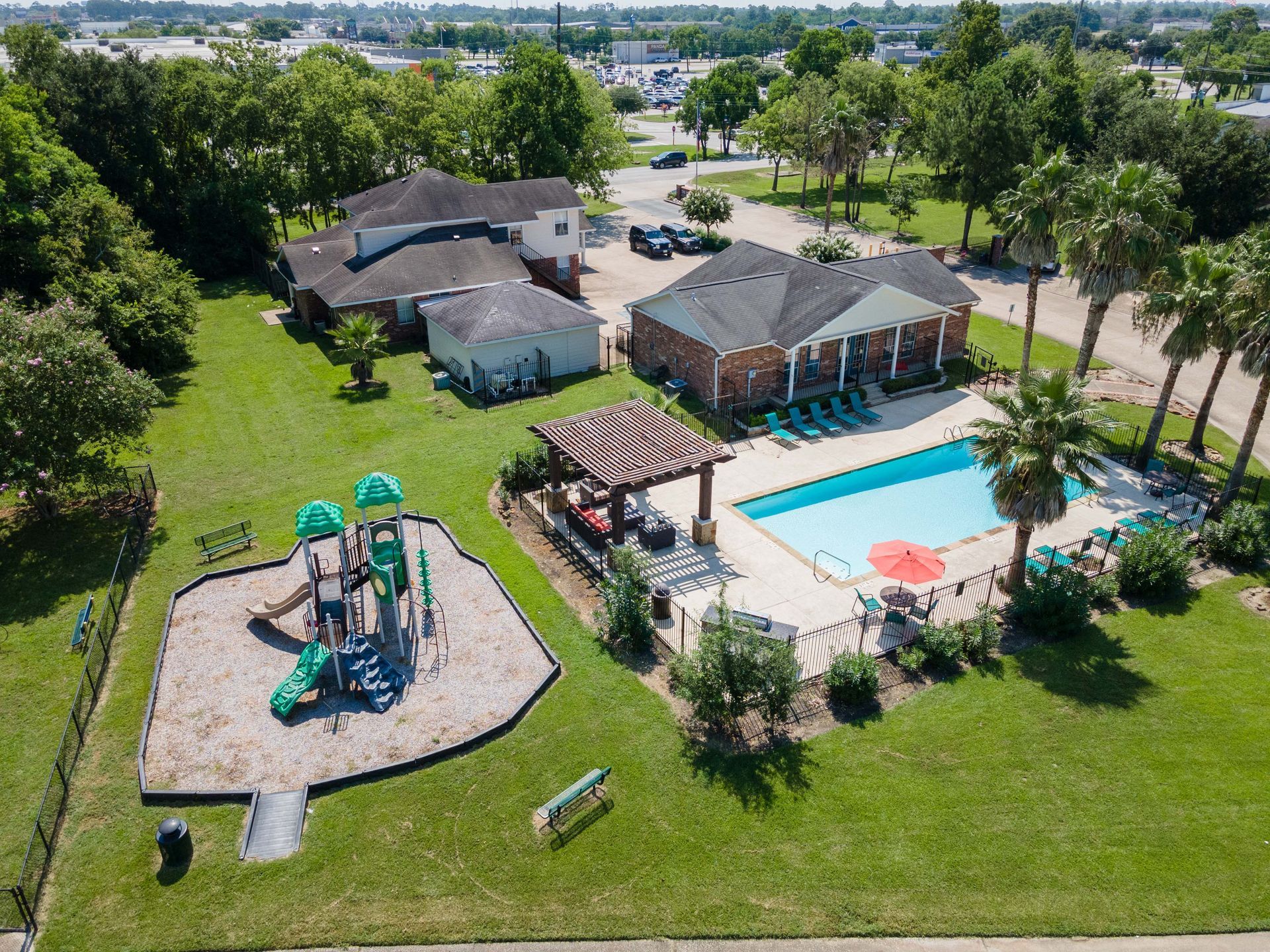 Aerial view of an apartment complex with a pool, playground, and trees on a sunny day.