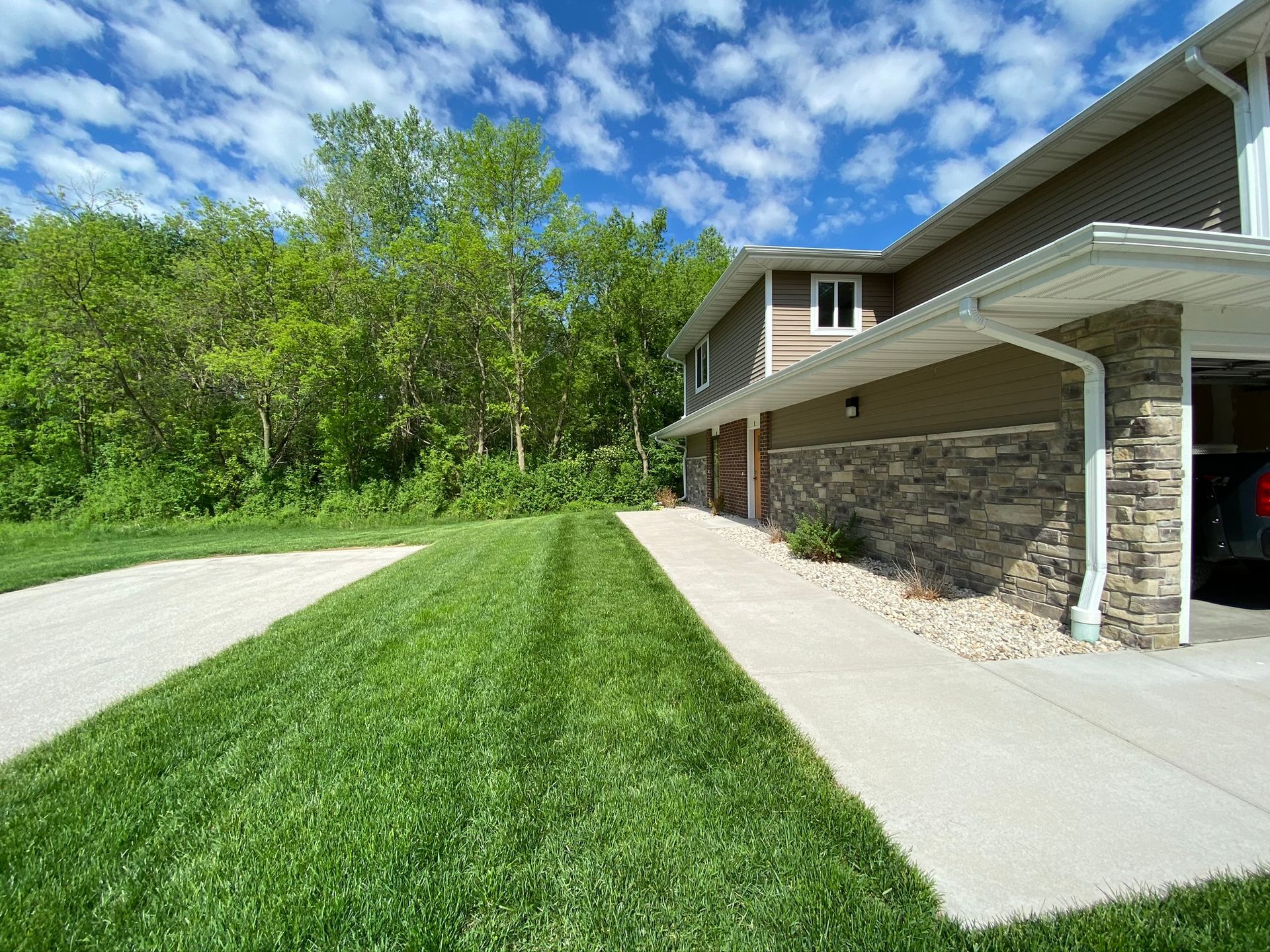 Suburban driveway beside a house with a carport, concrete walk, and green lawn under a blue sky