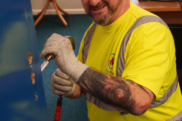 Man in yellow safety vest chiseling a blue surface; wearing gloves, visible tattoo.