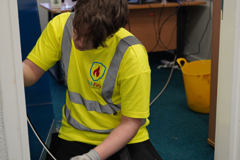 Person in a yellow safety vest working, possibly on wiring. Inside a room with a yellow bucket.