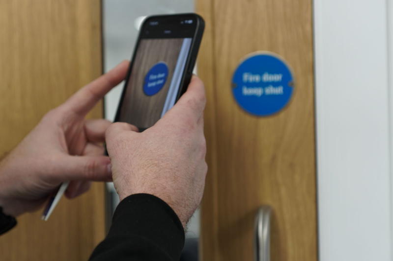 Person holding a phone, scanning a blue “Fire door keep shut” sign on a wooden door.