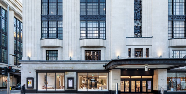Yellow brick building with white window frames and a flag in front, on a grassy hill under a blue sky.