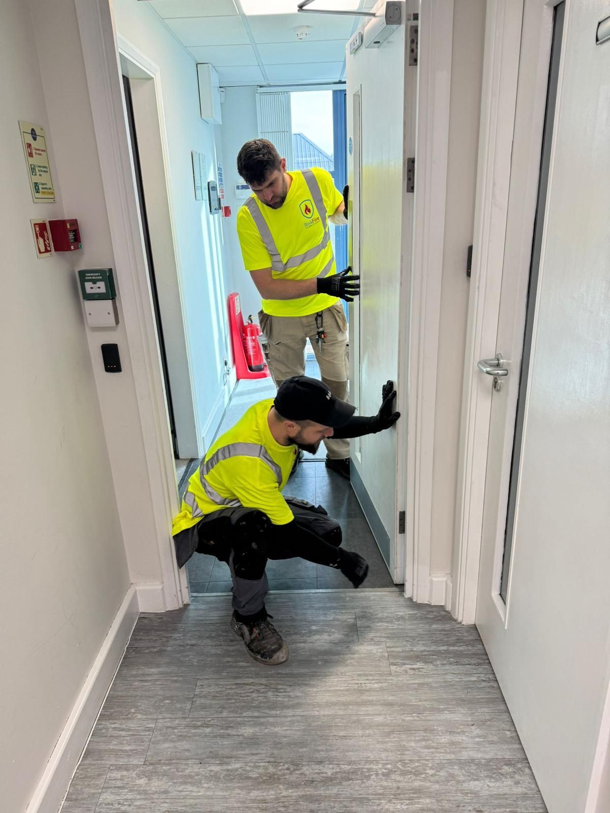 Two workers in high-vis vests install a white door in a hallway. One crouches, the other stands.