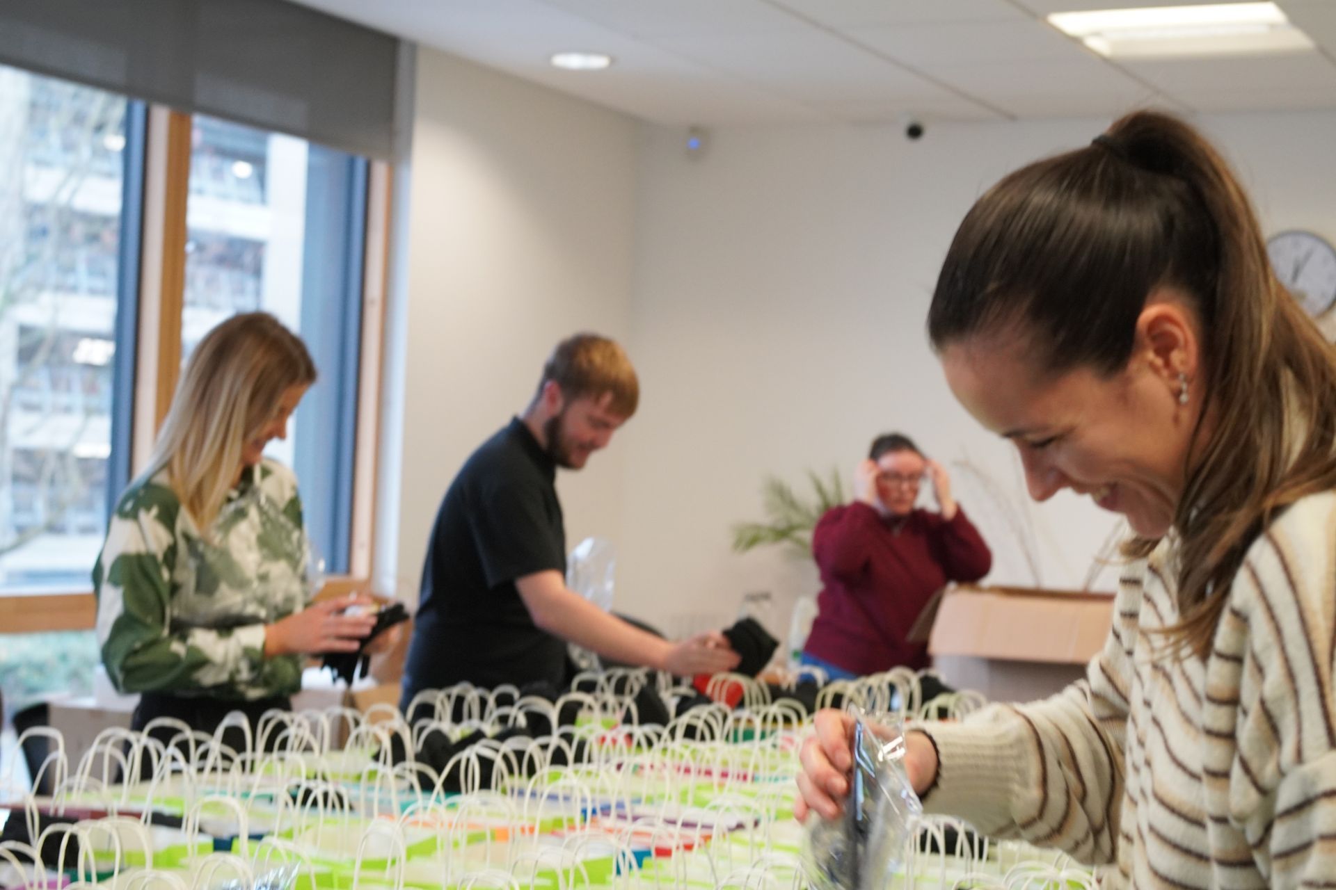 People packing items at a table in a brightly lit room. A woman smiles, others sort and pack.