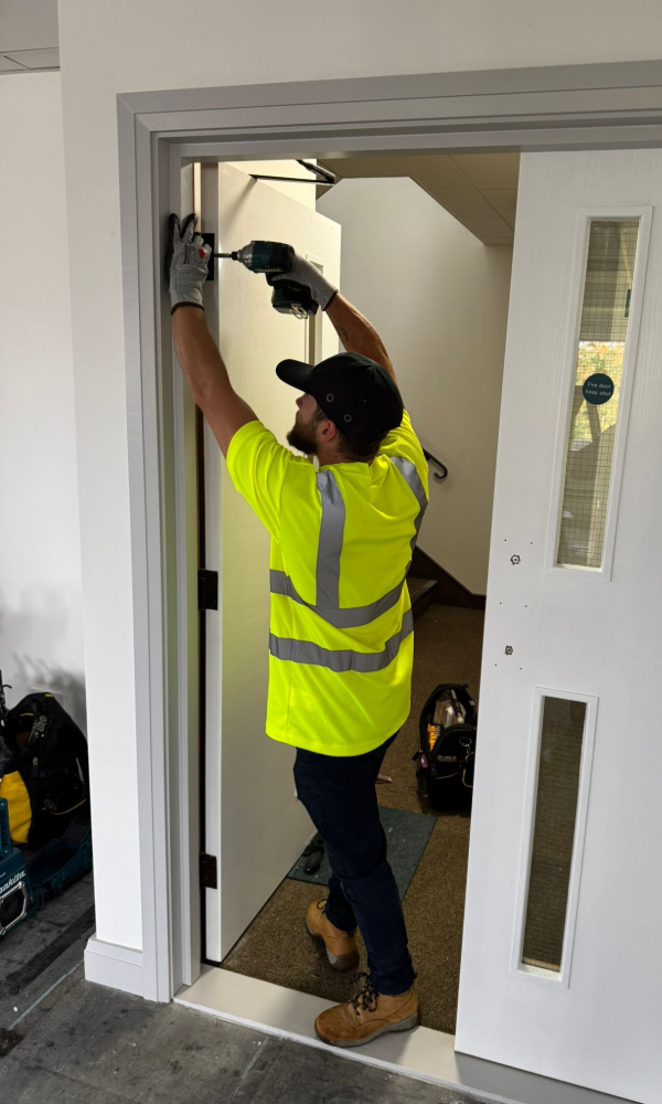 A person in a yellow safety vest installs a door closer on a white door frame with a power drill.