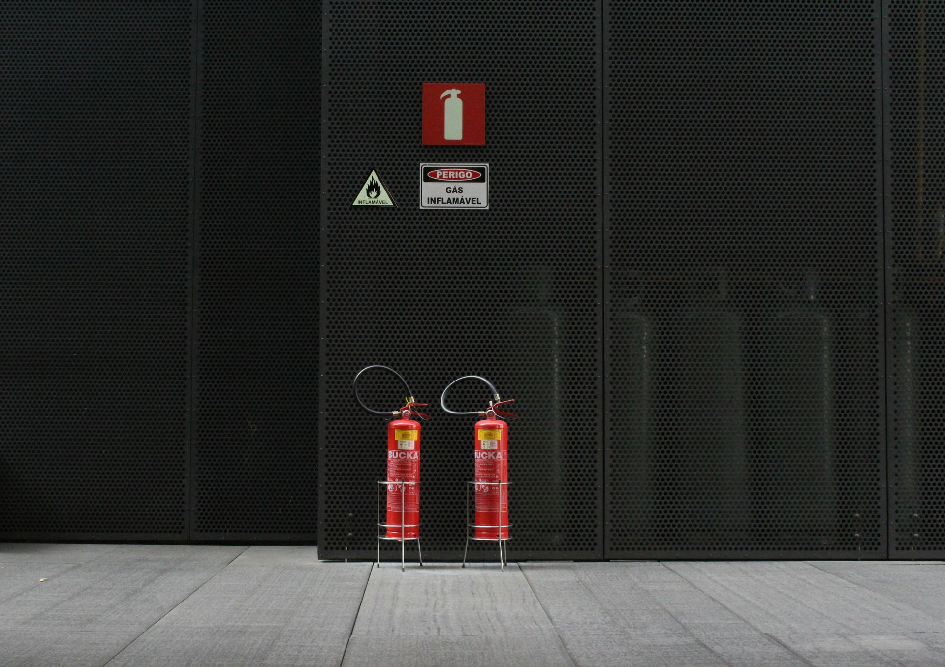 Two red fire extinguishers stand against a dark metal wall, with warning signs above.