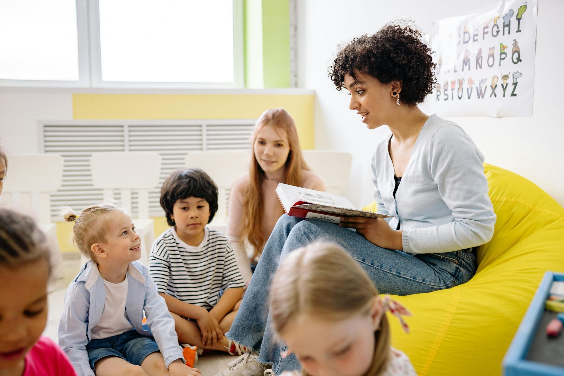 A woman is sitting on a bean bag chair reading a book to a group of children.