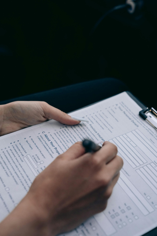 Person's hands holding a clipboard, writing on a form with a pen. Dark background.