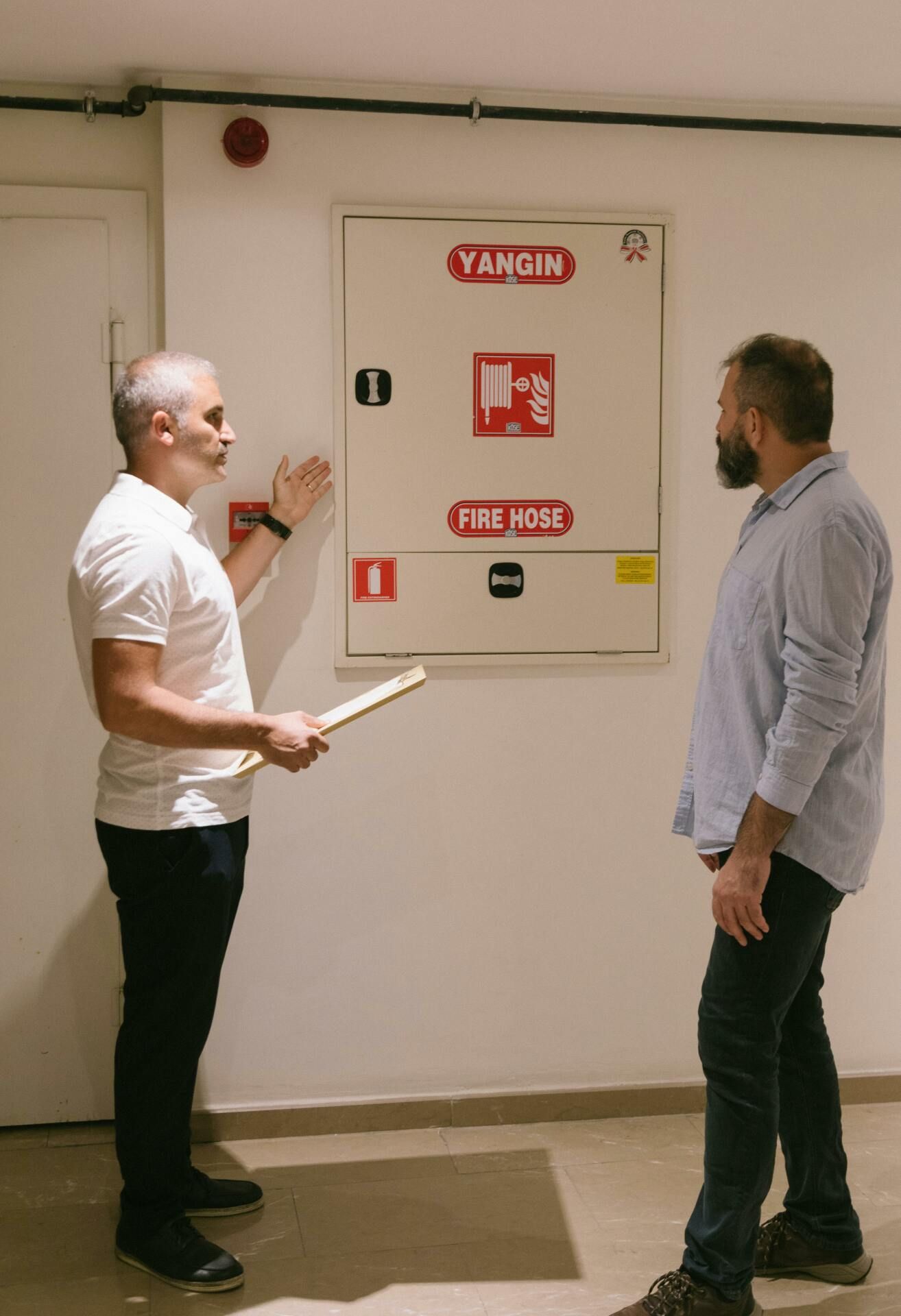 Two men are standing next to each other in front of a fire extinguisher.