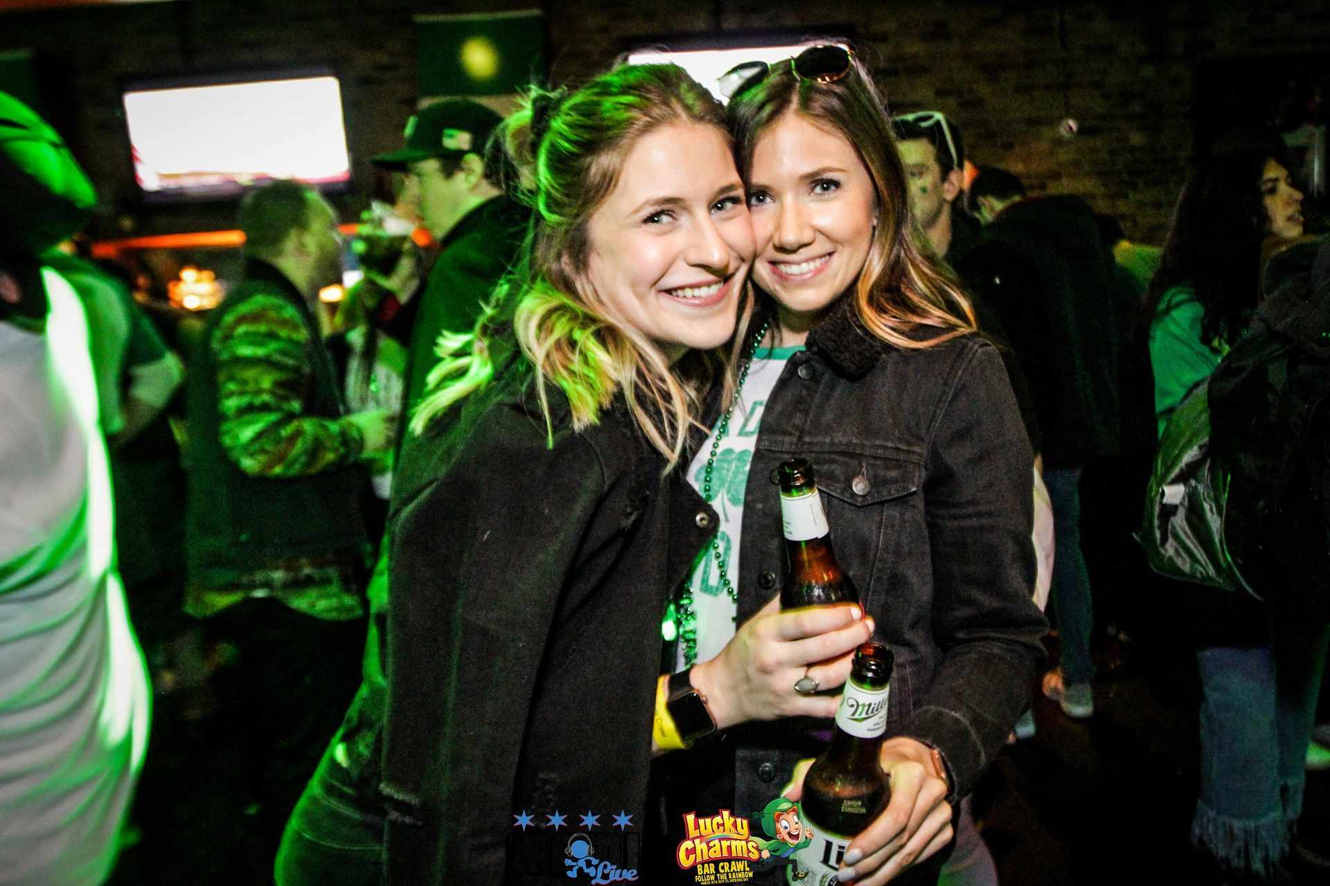 Two women are posing for a picture at a party while holding beer bottles.