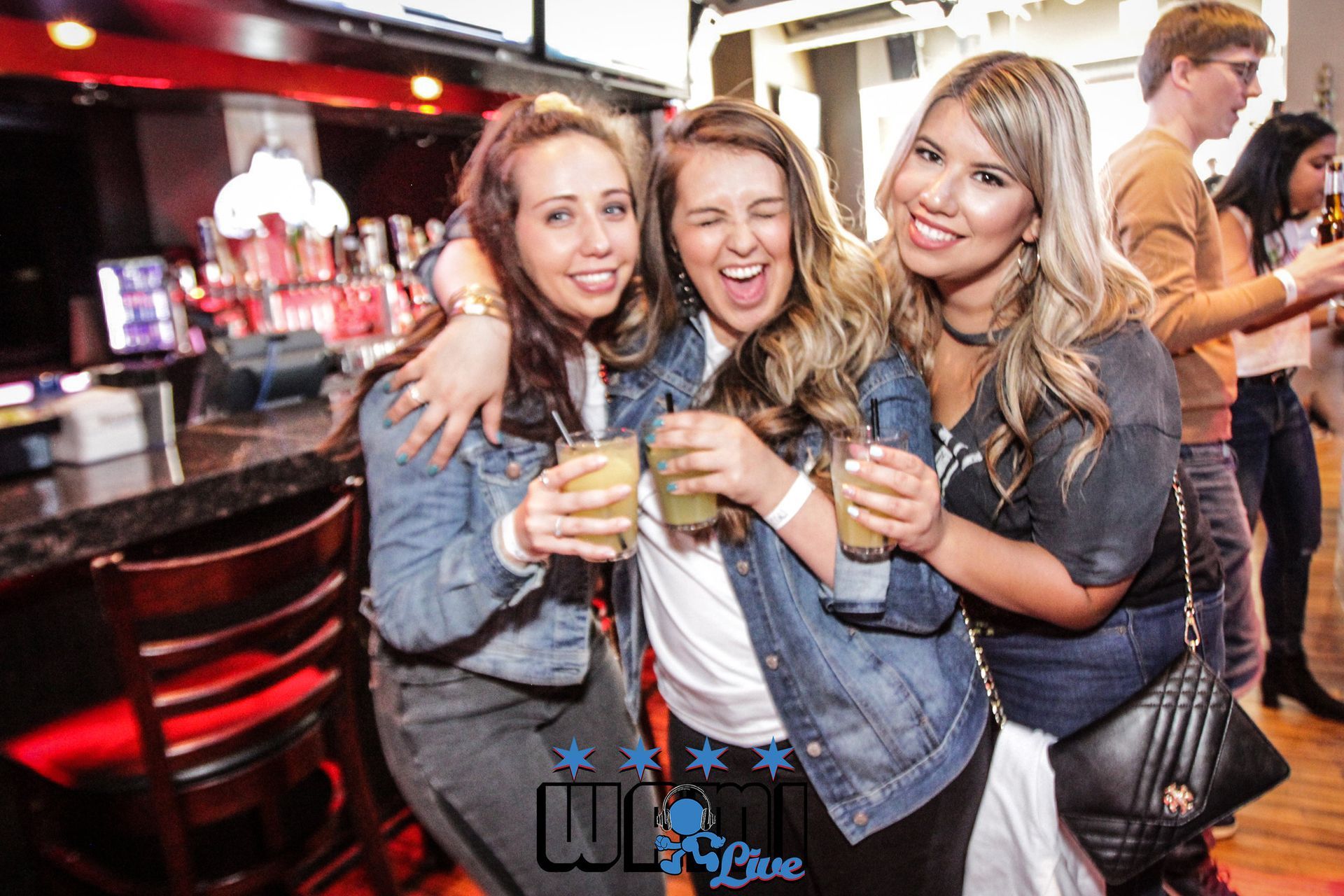 Three women are posing for a picture in a bar while holding drinks.