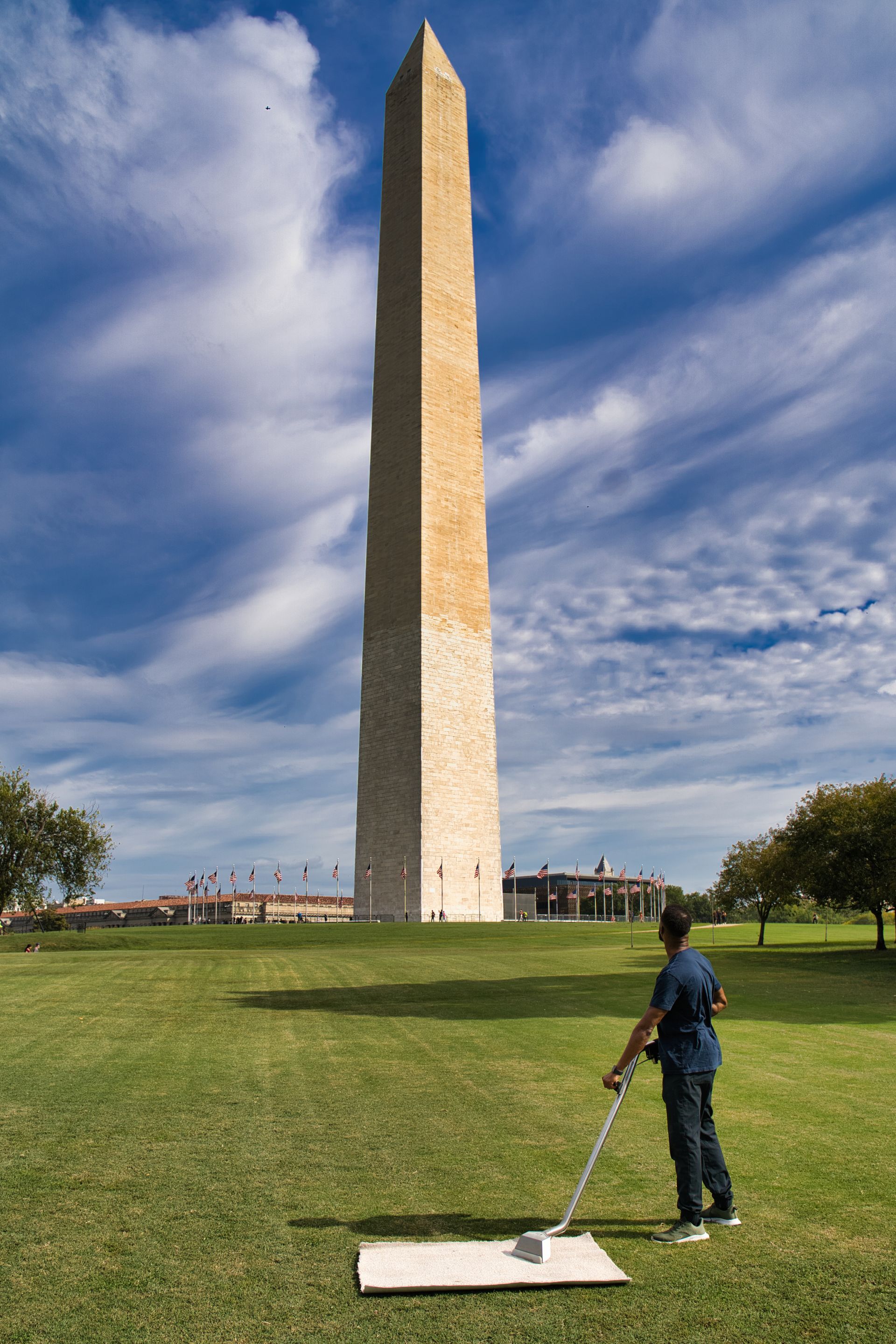 Man uses a metal detector on the grass in front of the Washington Monument on a sunny day.