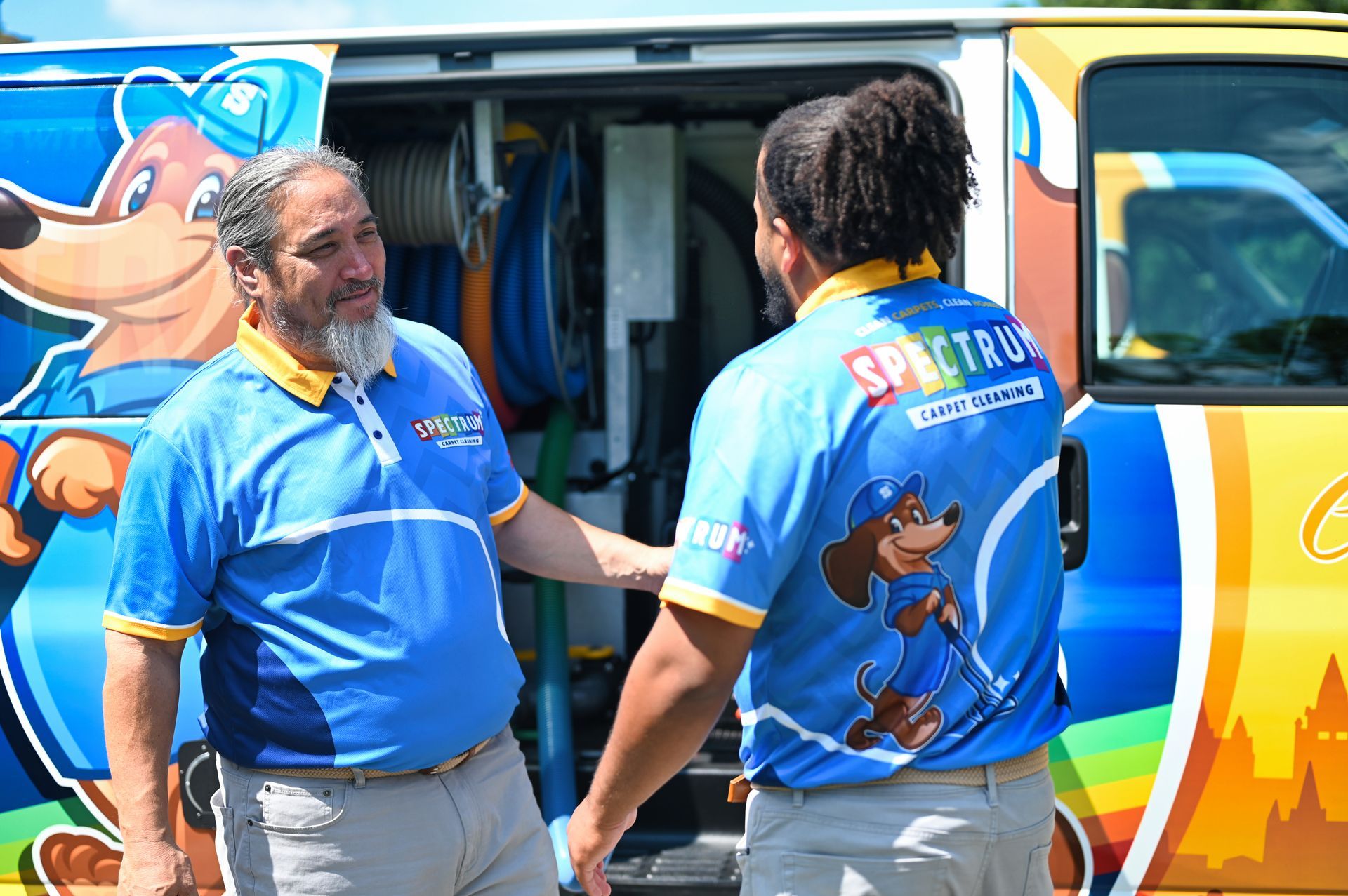 Two Spectrum carpet cleaners, one gray-bearded, talking in front of their van with cartoon dog logo.