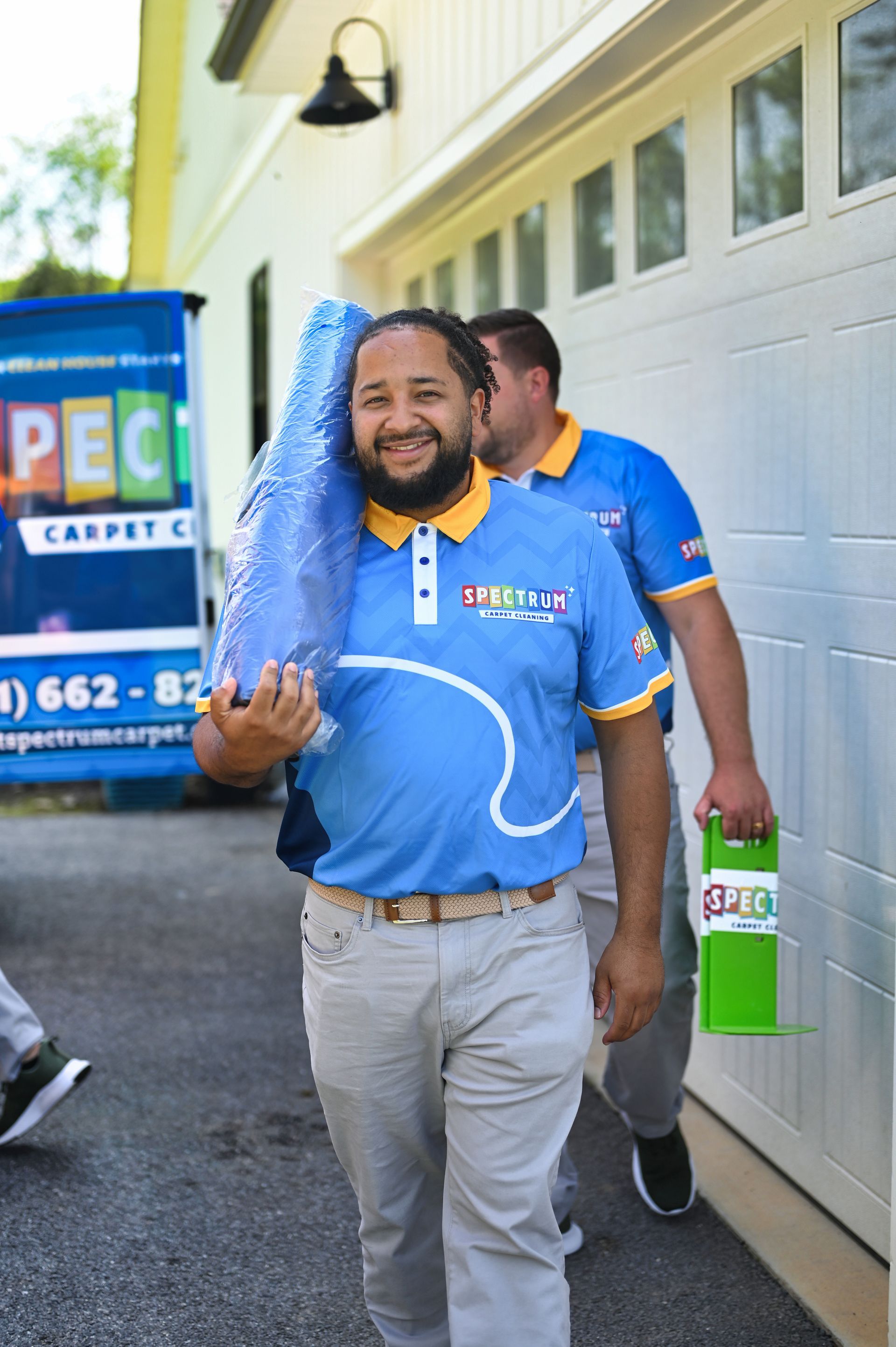 Man in blue shirt carries equipment, smiling. Another follows. Outside a garage.