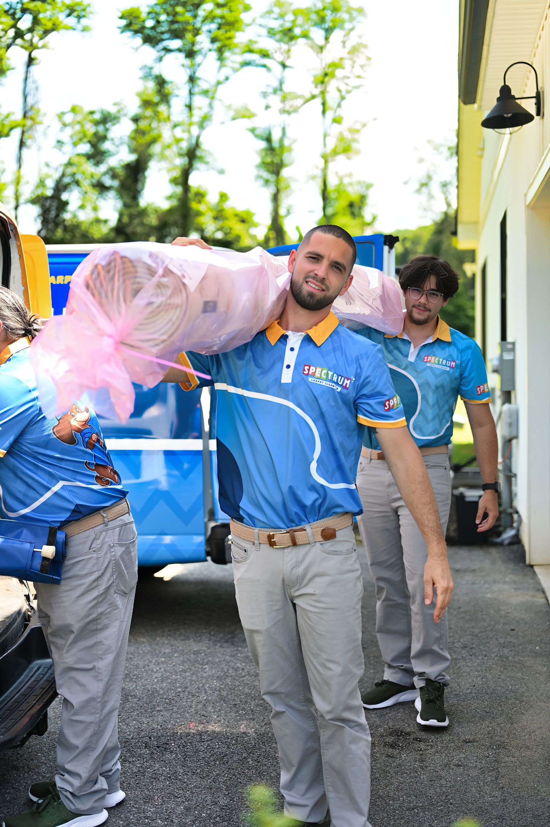Three men in blue shirts carry a rolled rug, exiting a truck. Outdoors, light colored building behind.