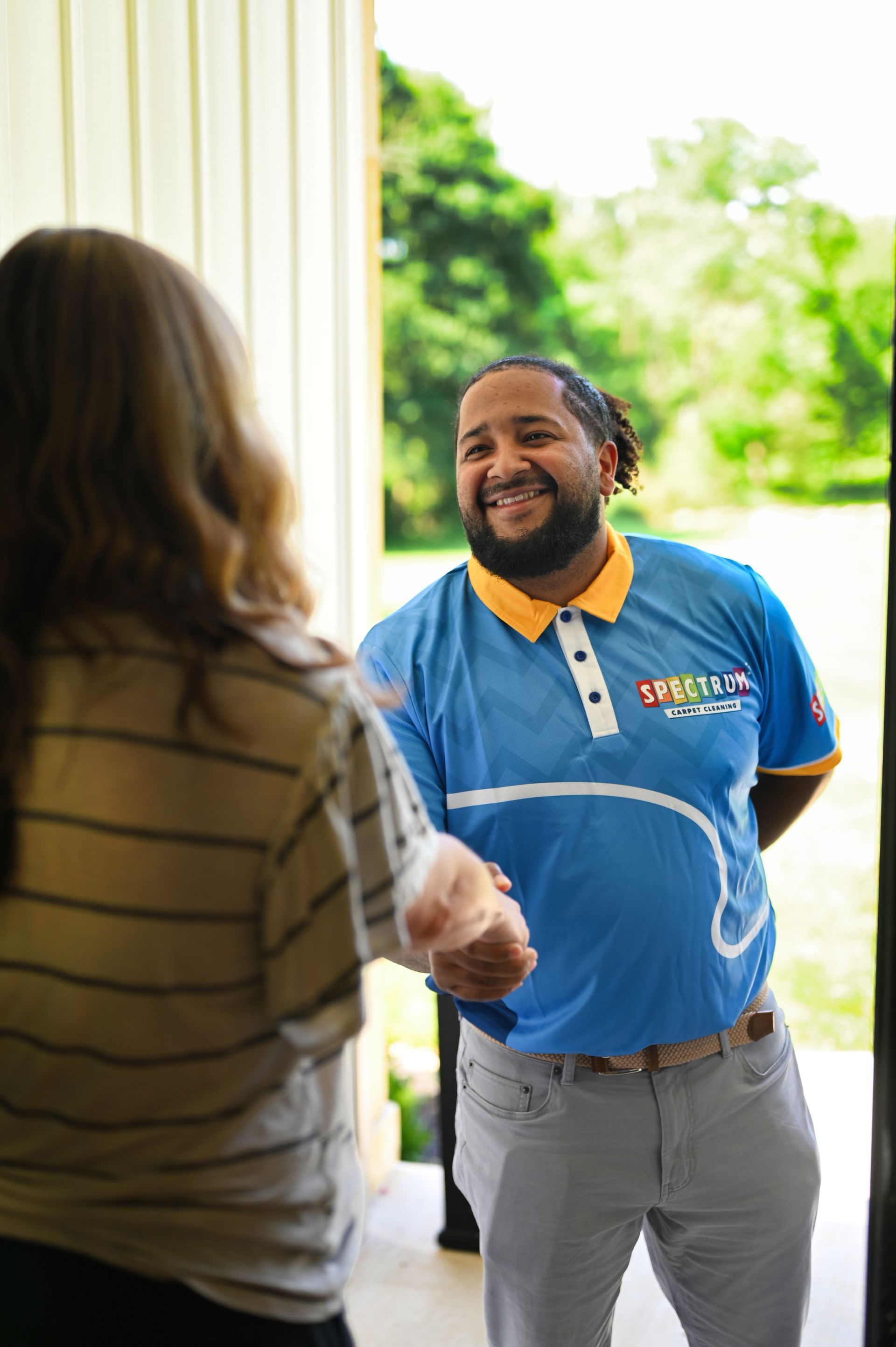 Man in blue shirt greets a person at a doorway, smiling. He has dreadlocks. Green background.