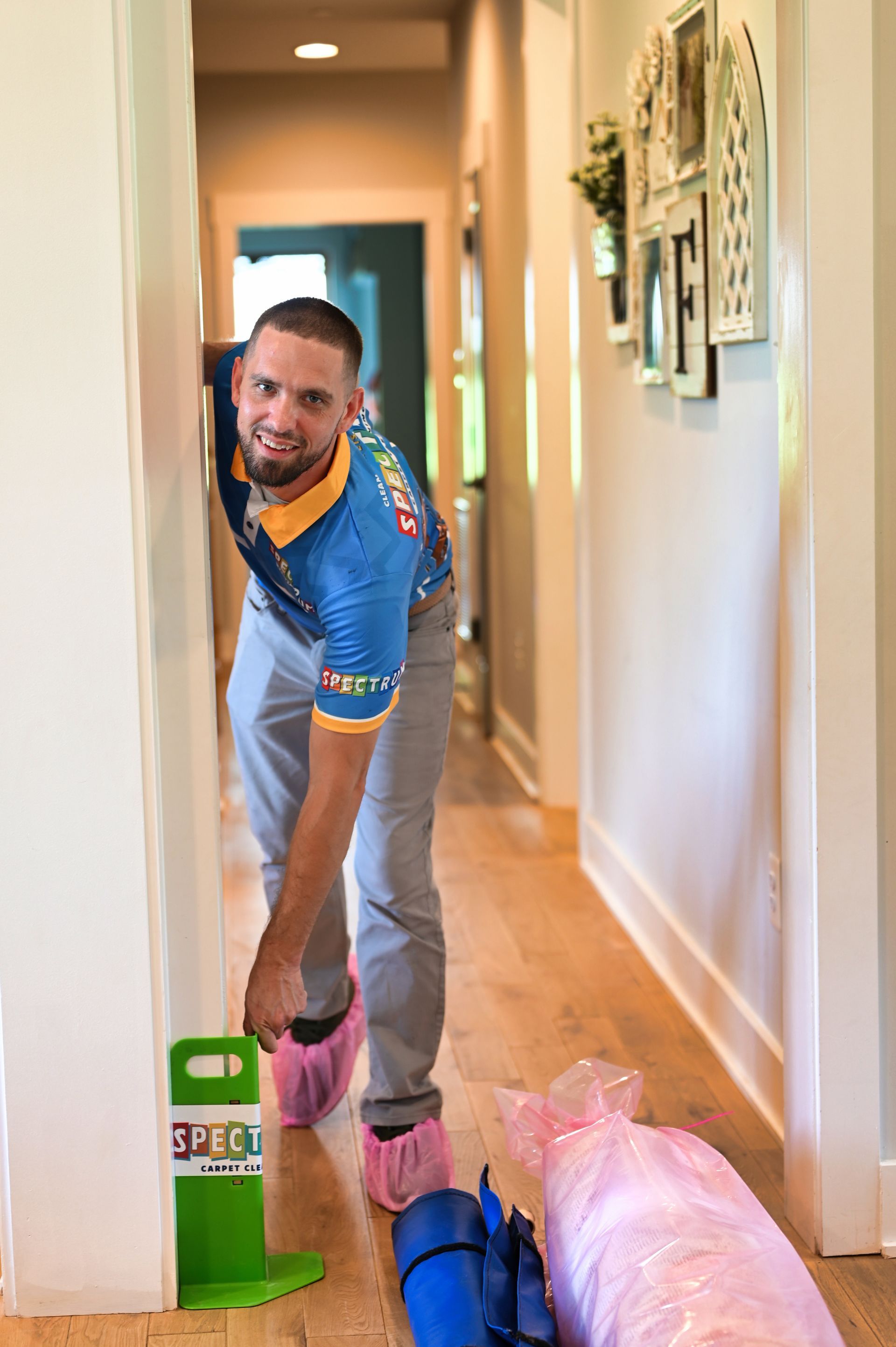 Man in blue shirt and grey pants kneels in a hallway, wearing pink shoe covers.  Hallway with art and equipment.