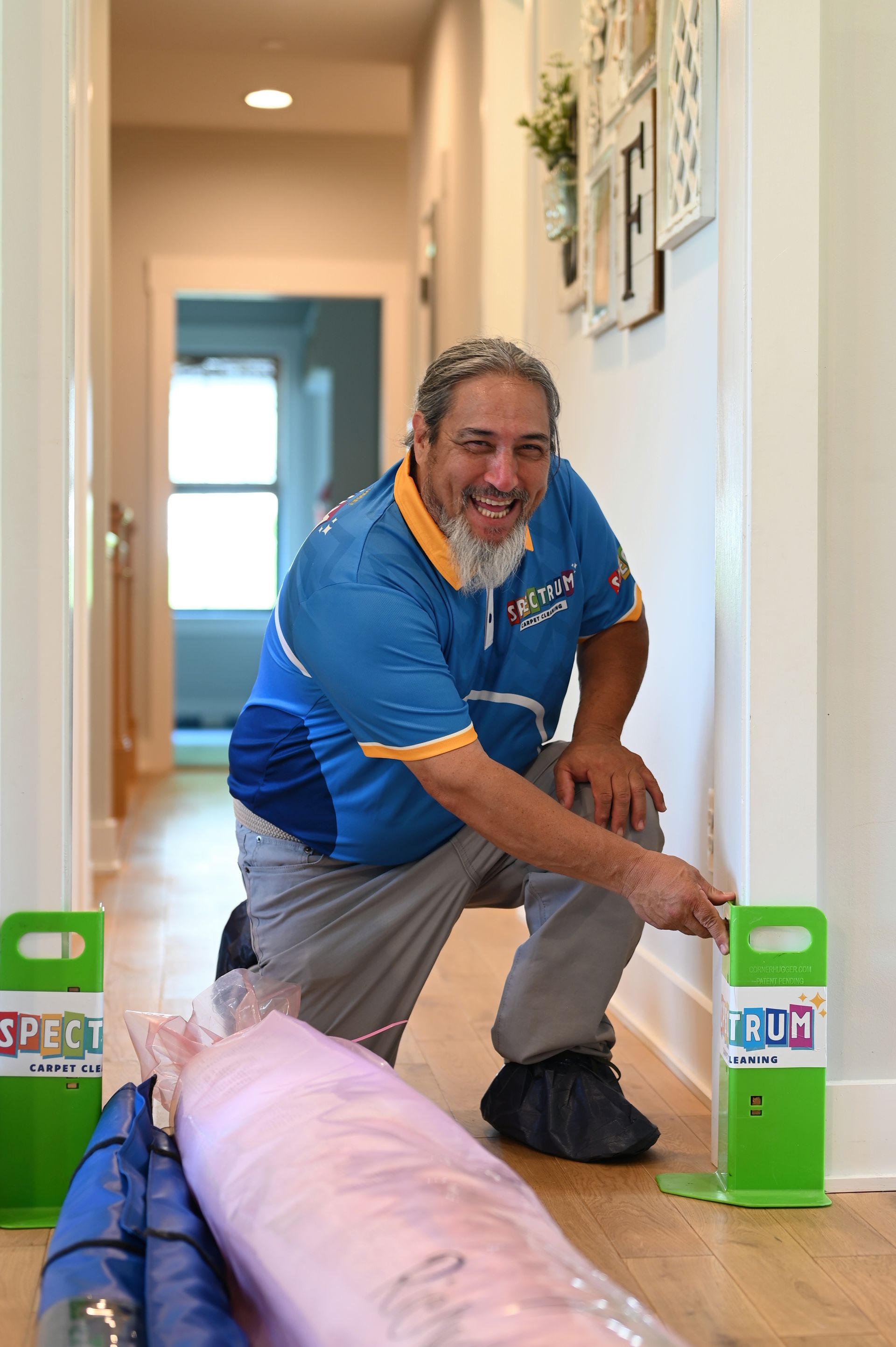 Man kneels in hallway, pointing at wall. He wears a blue shirt, smiling. Green supports and wrapped items are near him.