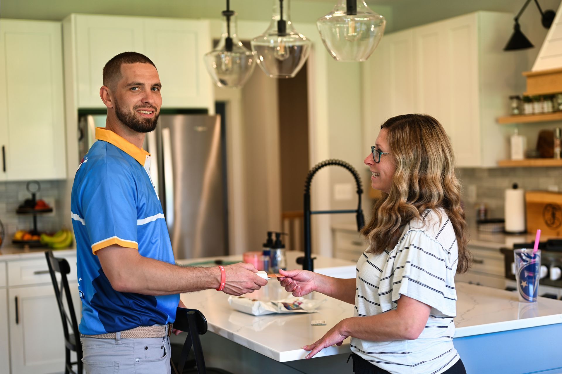 Man in blue shirt hands something to woman in kitchen. White cabinets, island, and pendant lights.