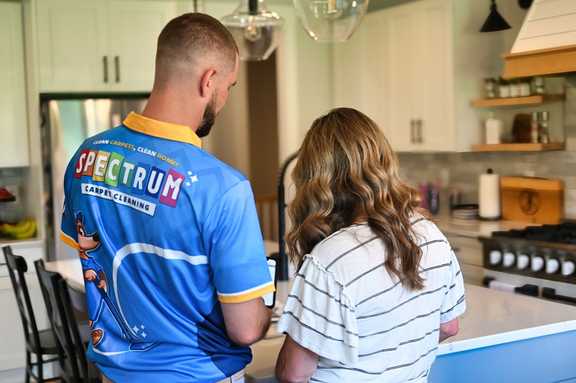 Man in Spectrum shirt and woman in a kitchen, looking at something near the sink.