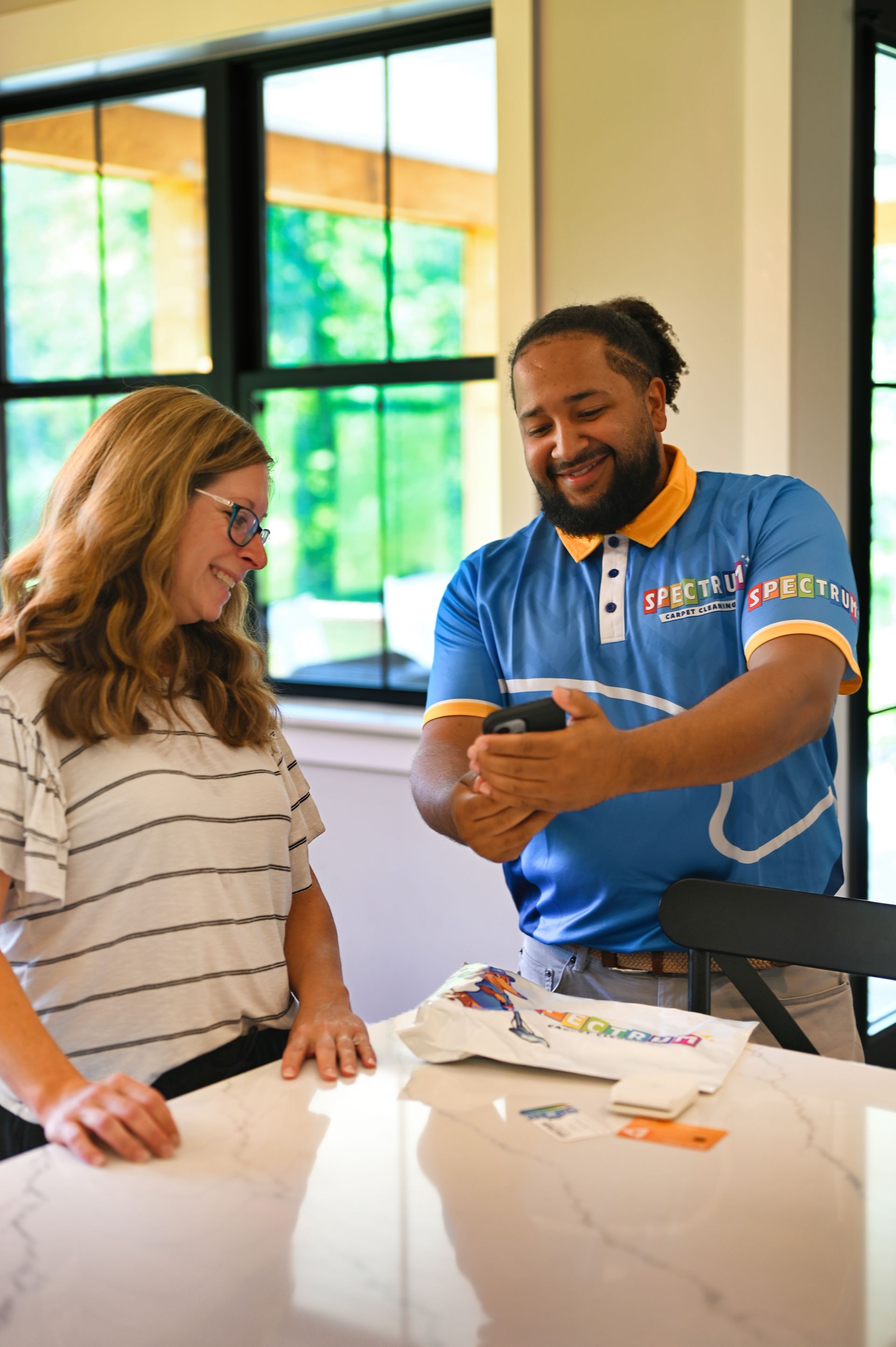 Woman and man looking at phone, smiling. Man wearing blue shirt, standing in bright kitchen.