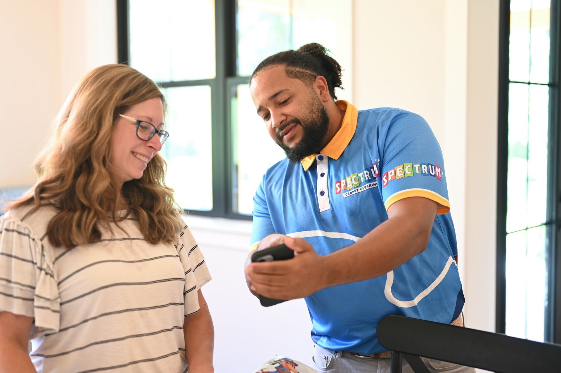 Woman and man looking at a phone. Man in blue polo shirt, woman wearing glasses. Indoors, near a window.