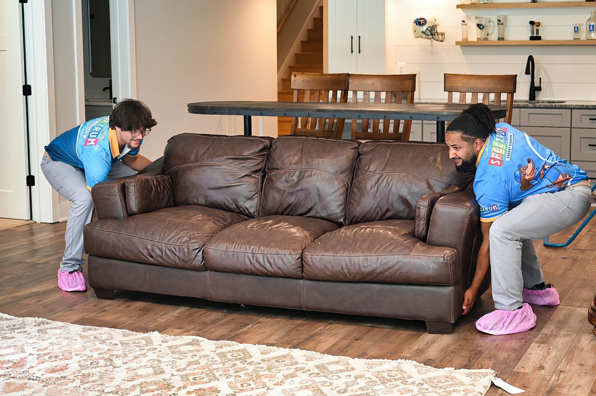 Two movers in blue shirts and pink shoe covers lift a brown leather couch in a home.