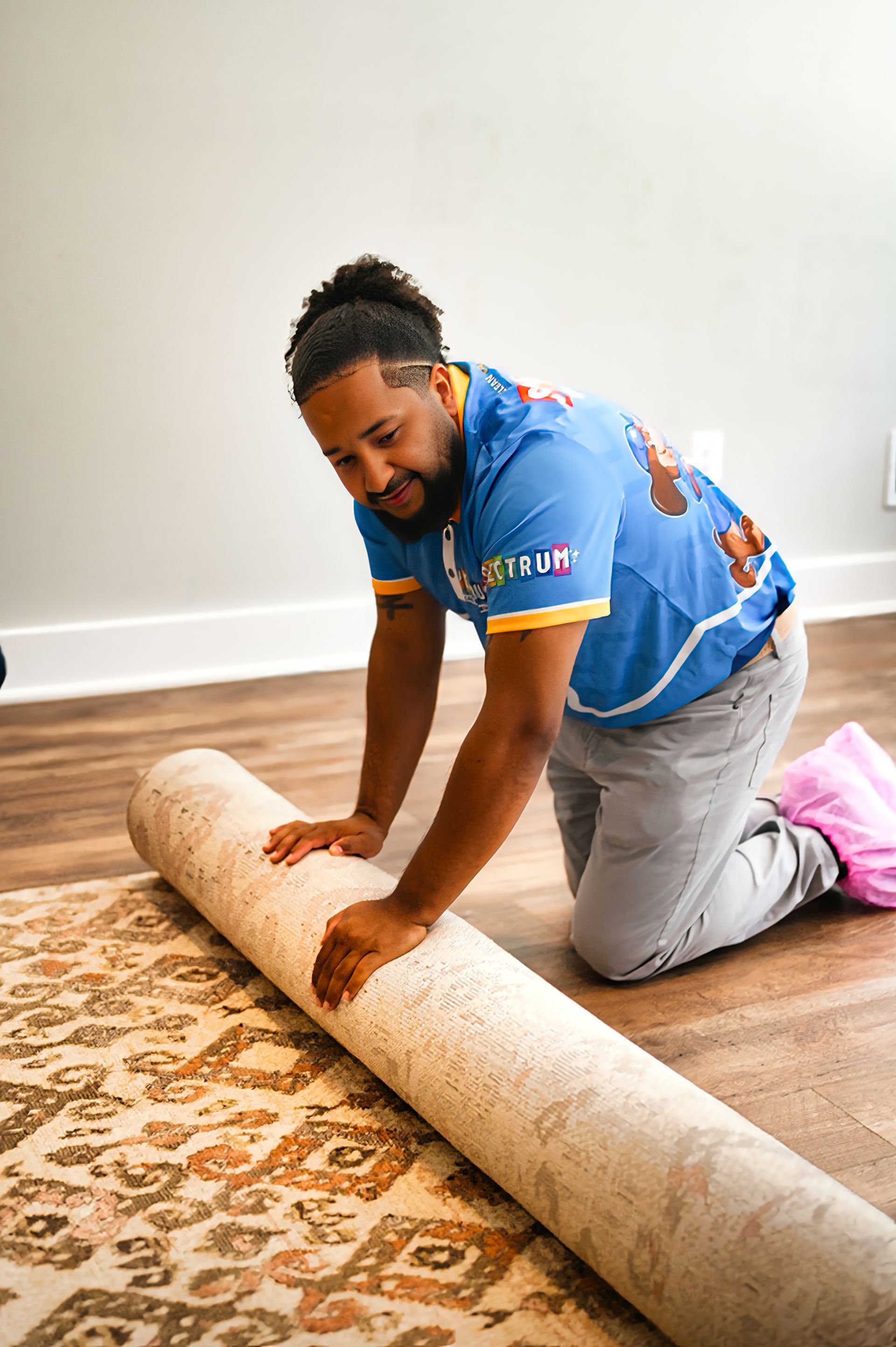 Man kneeling, rolling out a patterned rug on a hardwood floor. He's wearing a blue shirt and gray pants.