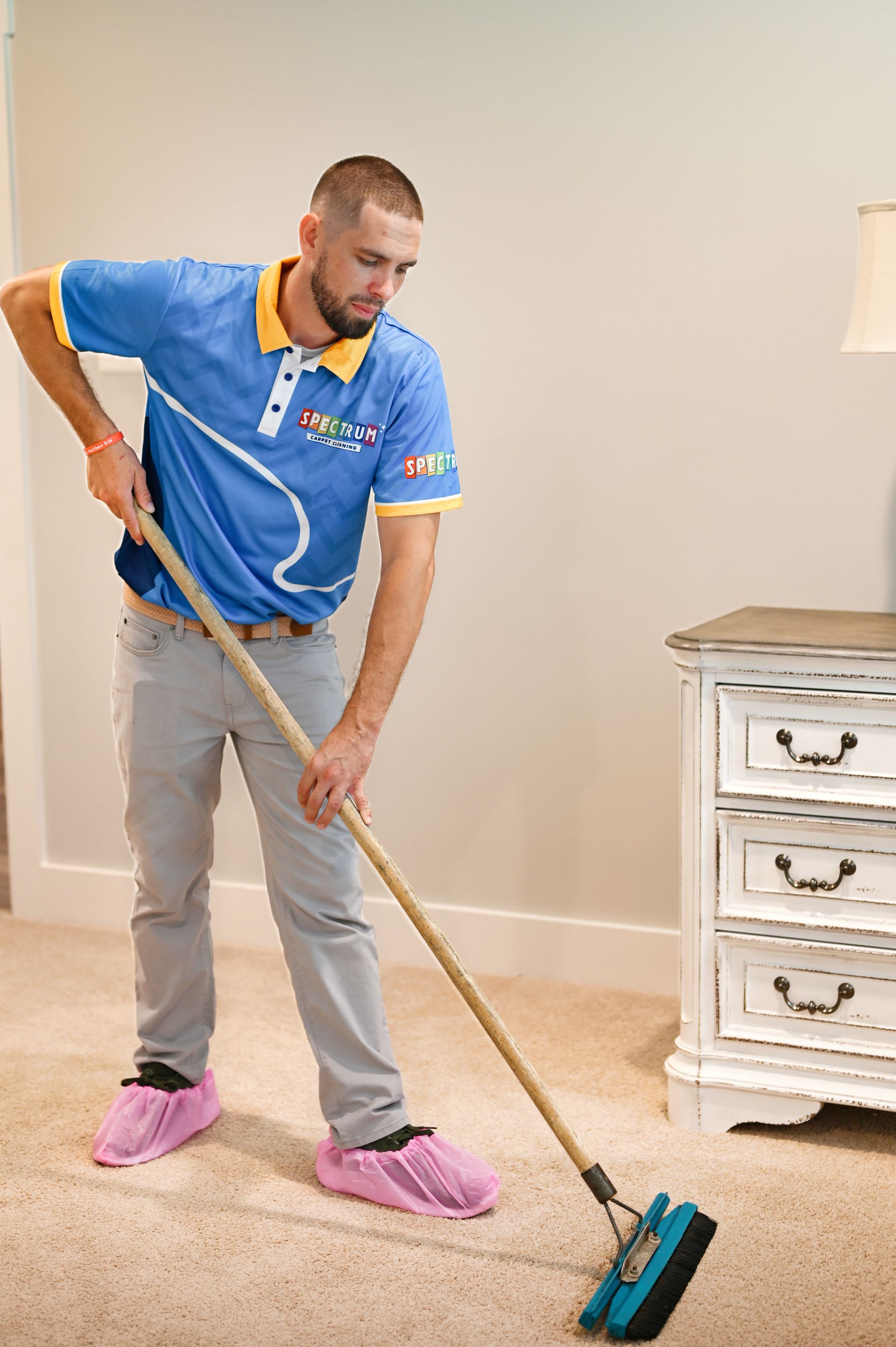 Man in blue shirt, light pants, and pink shoe covers sweeping carpet near a dresser in a room.