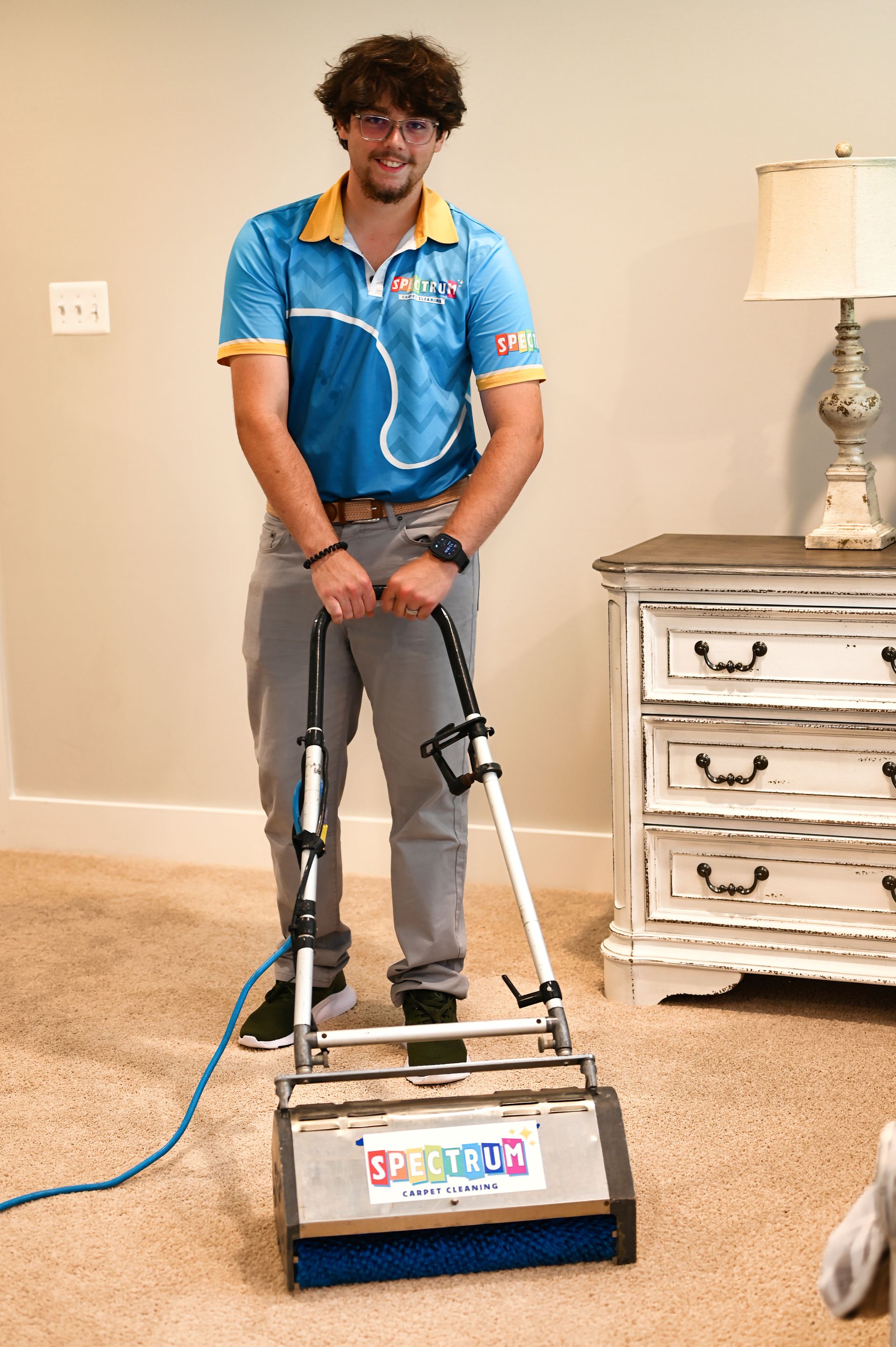 Man with curly hair cleaning carpet with a machine in a room with a nightstand.