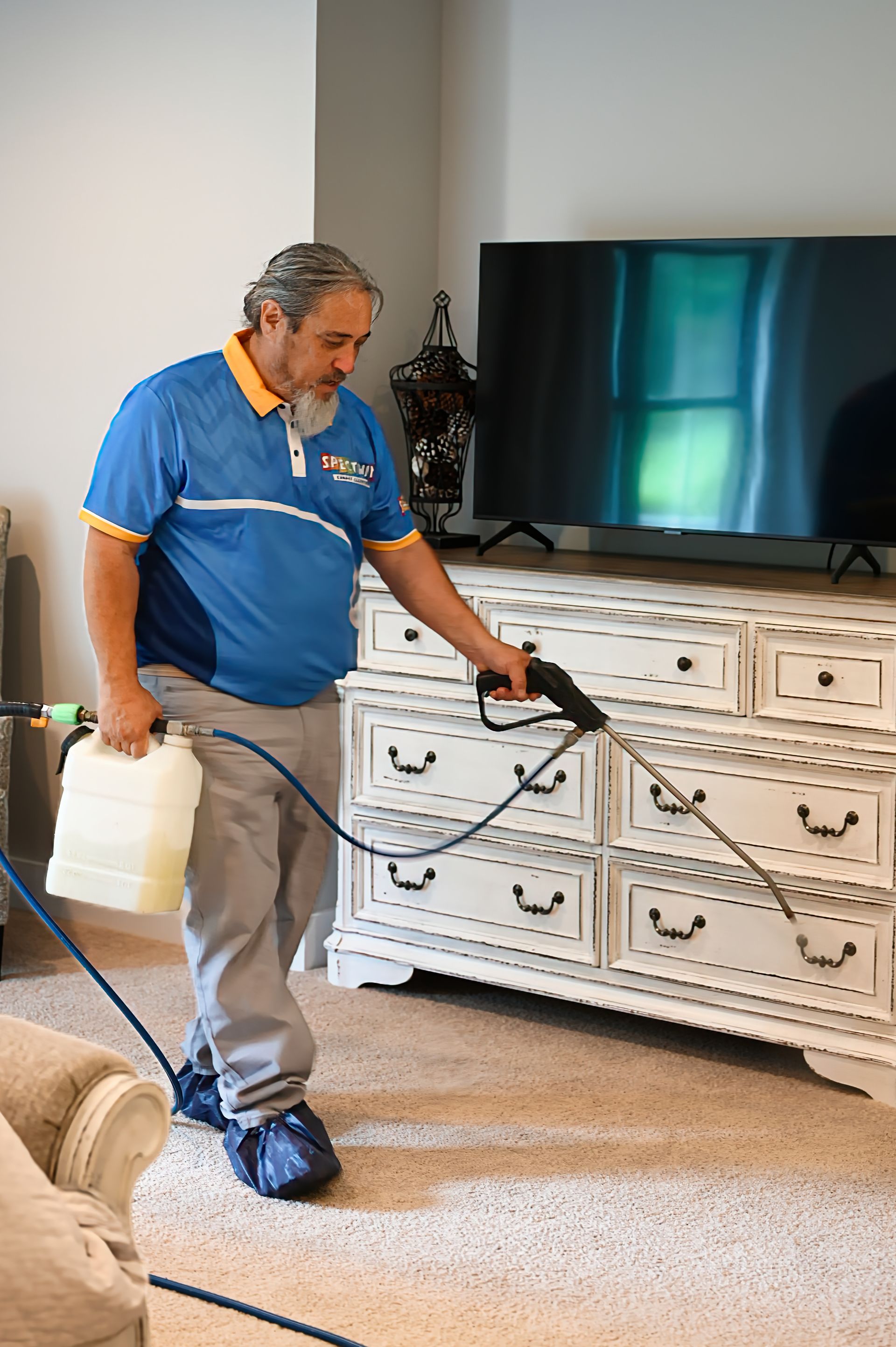 Man in blue shirt spraying carpet near a white dresser and TV; in a living room.