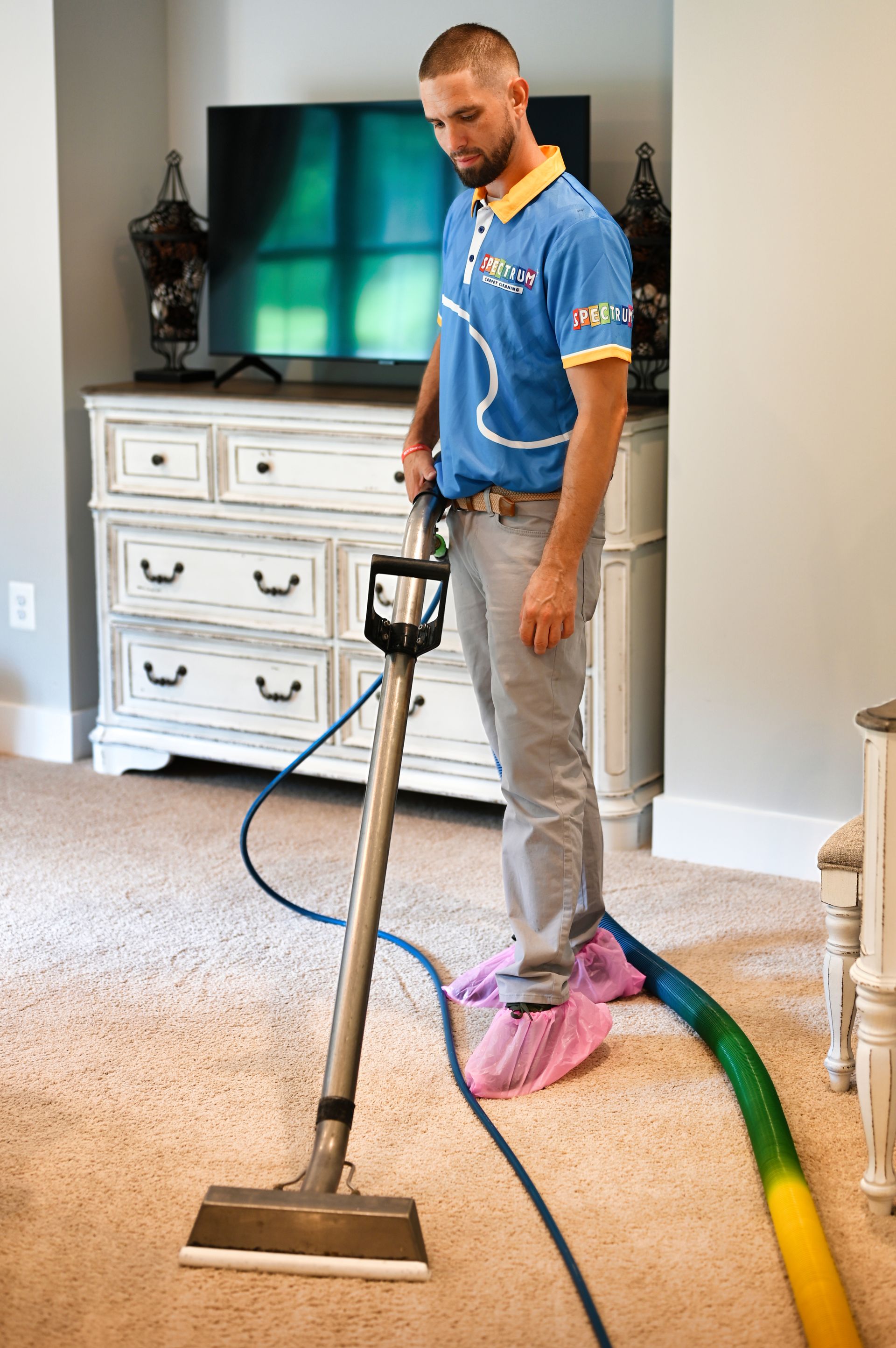 Man cleaning a carpet with a machine; wearing a blue shirt and pink shoe covers, in a bedroom.