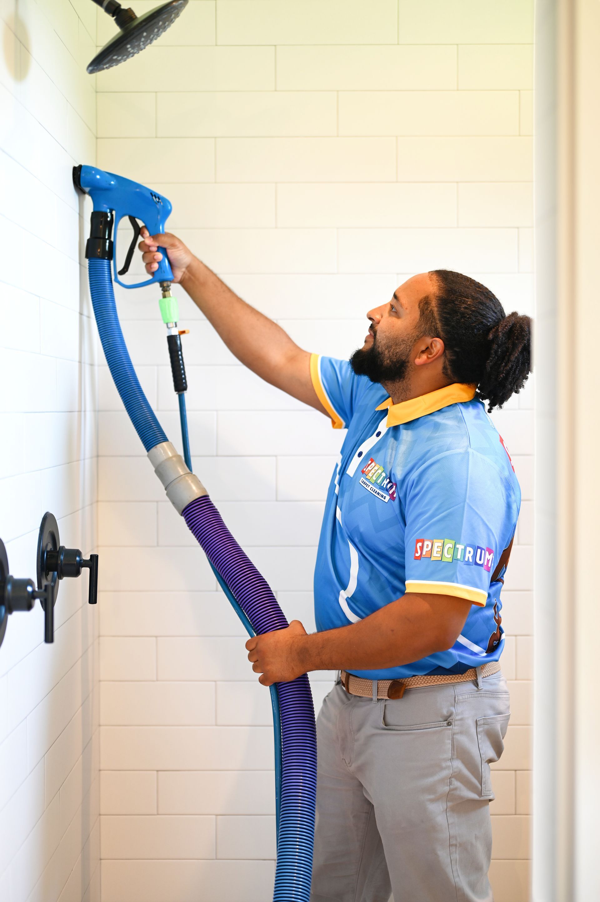 Man cleaning shower with a blue and purple hose, white tiled wall.