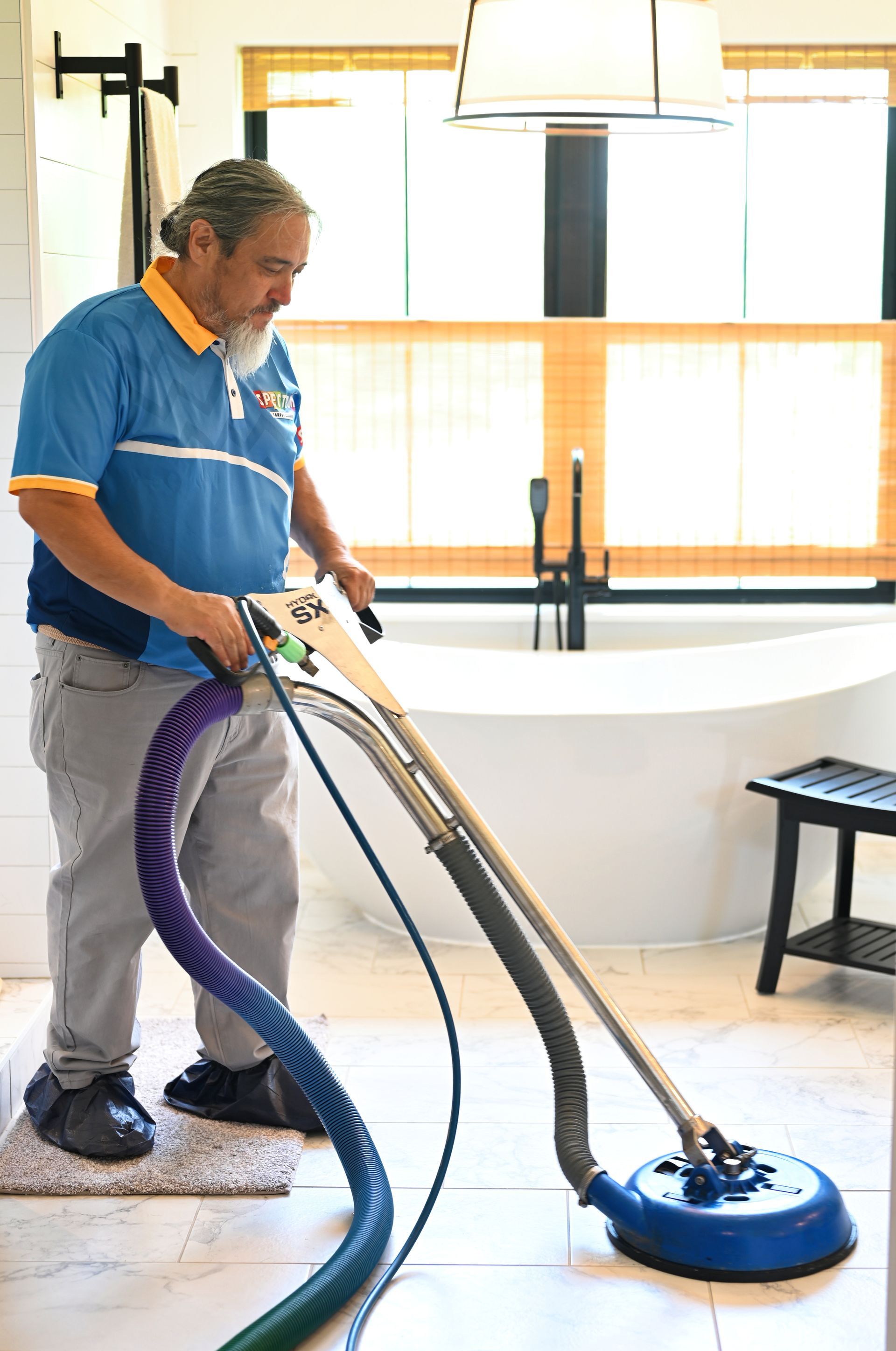 Man cleaning bathroom floor with a machine. The room has a tub and window.
