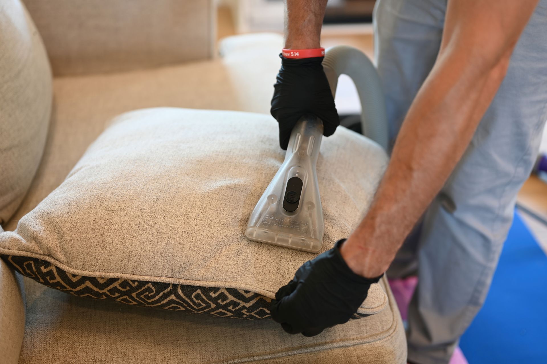 Person wearing black gloves cleaning a beige couch cushion with a vacuum cleaner attachment.