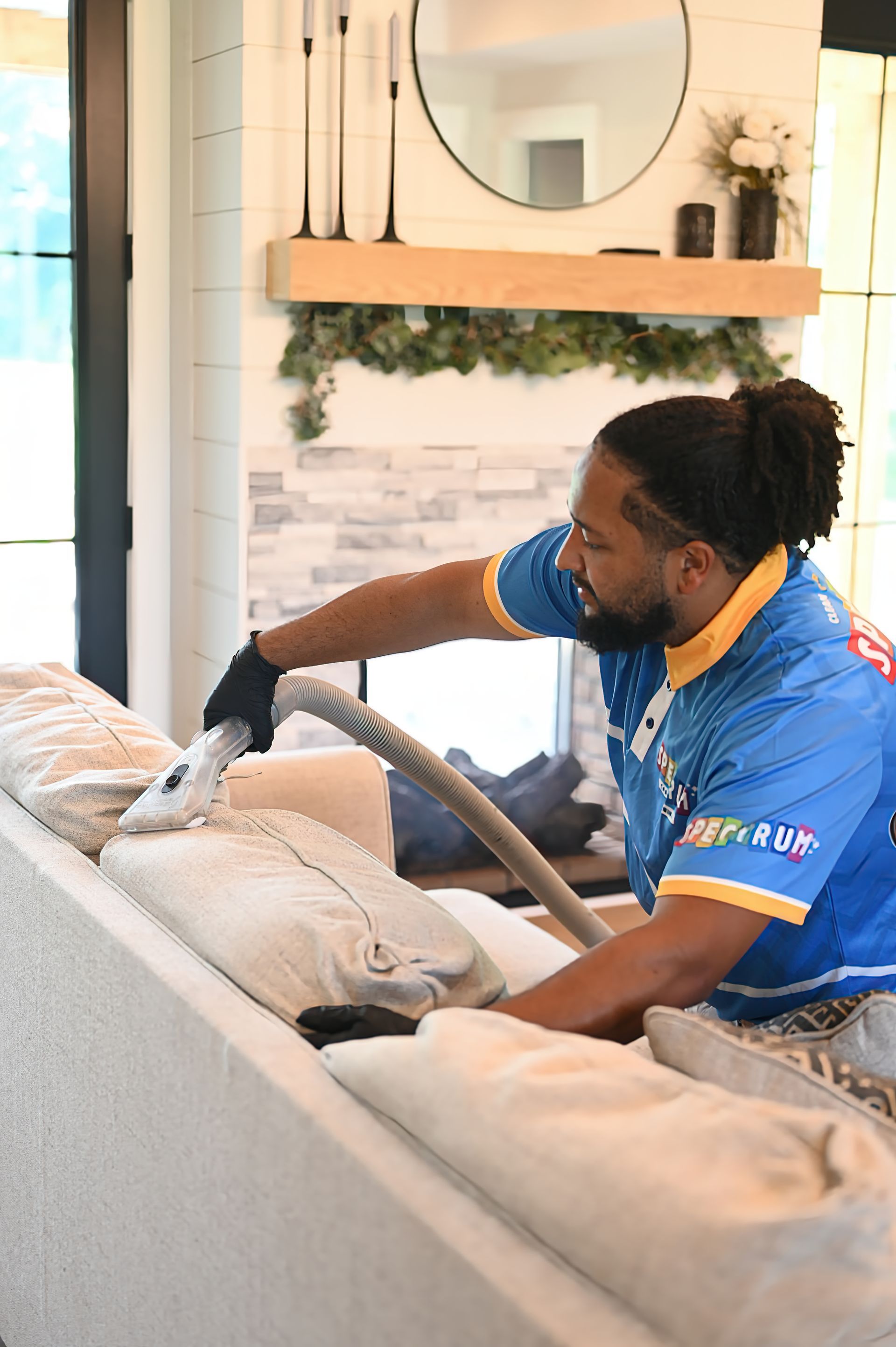 Man in blue shirt vacuums a light-colored couch in a home setting.