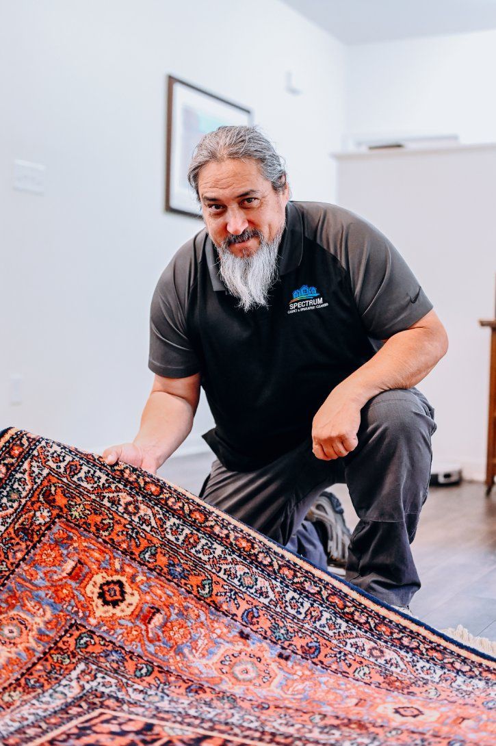 Man kneeling, inspecting a patterned rug. He's in a room, wearing work clothes.