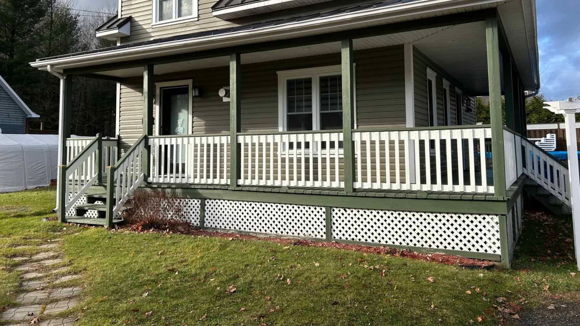Une maison avec un porche, un bardage vert et une balustrade blanche. Un chemin de pierre traverse la cour.
