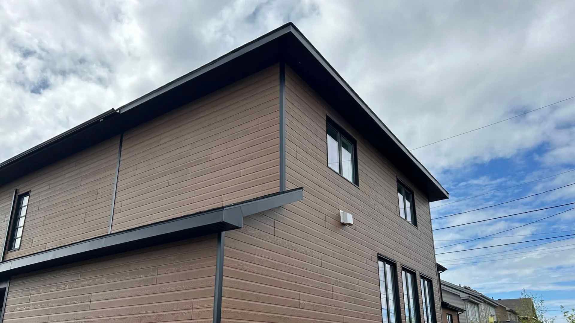 Maison brune à deux étages avec des boiseries noires sous un ciel nuageux.