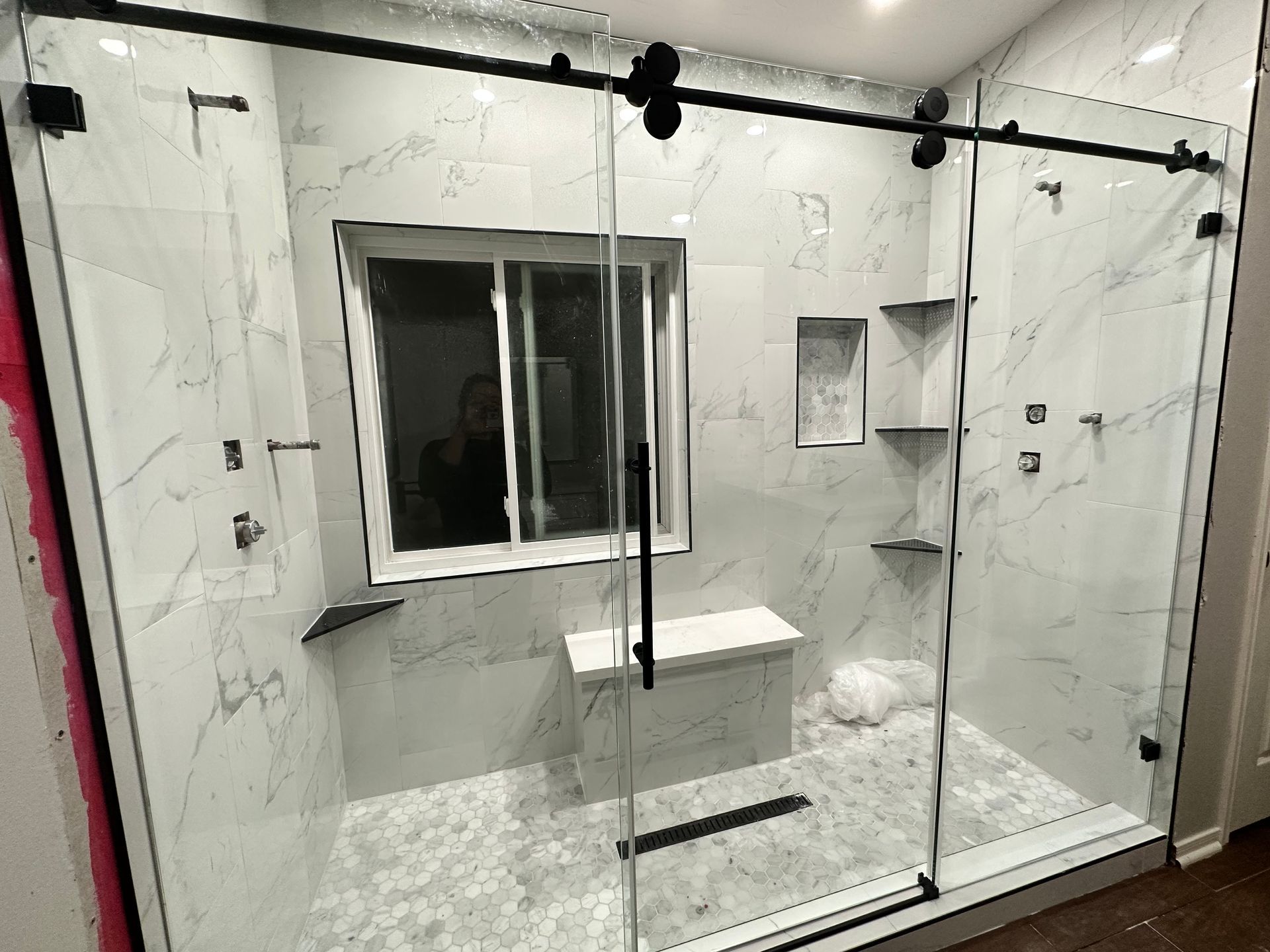 Modern glass-enclosed shower with marble-look tile, built-in bench, hexagon tile floor, and matte black hardware by A&J Remodel and Construction