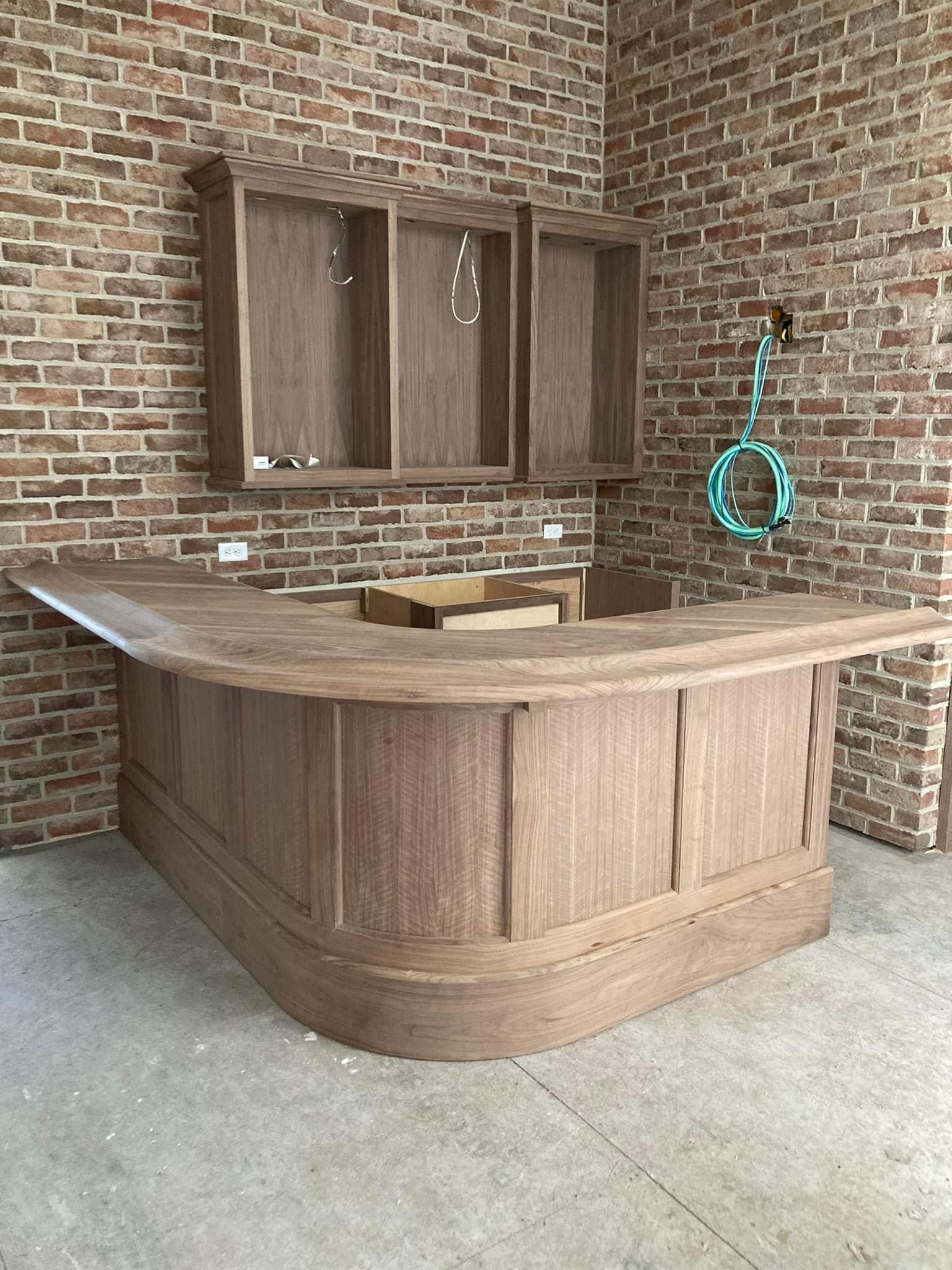 Custom-built indoor bar with curved wood paneling and matching upper cabinets, installed by A&J Remodel and Construction against a brick accent wall