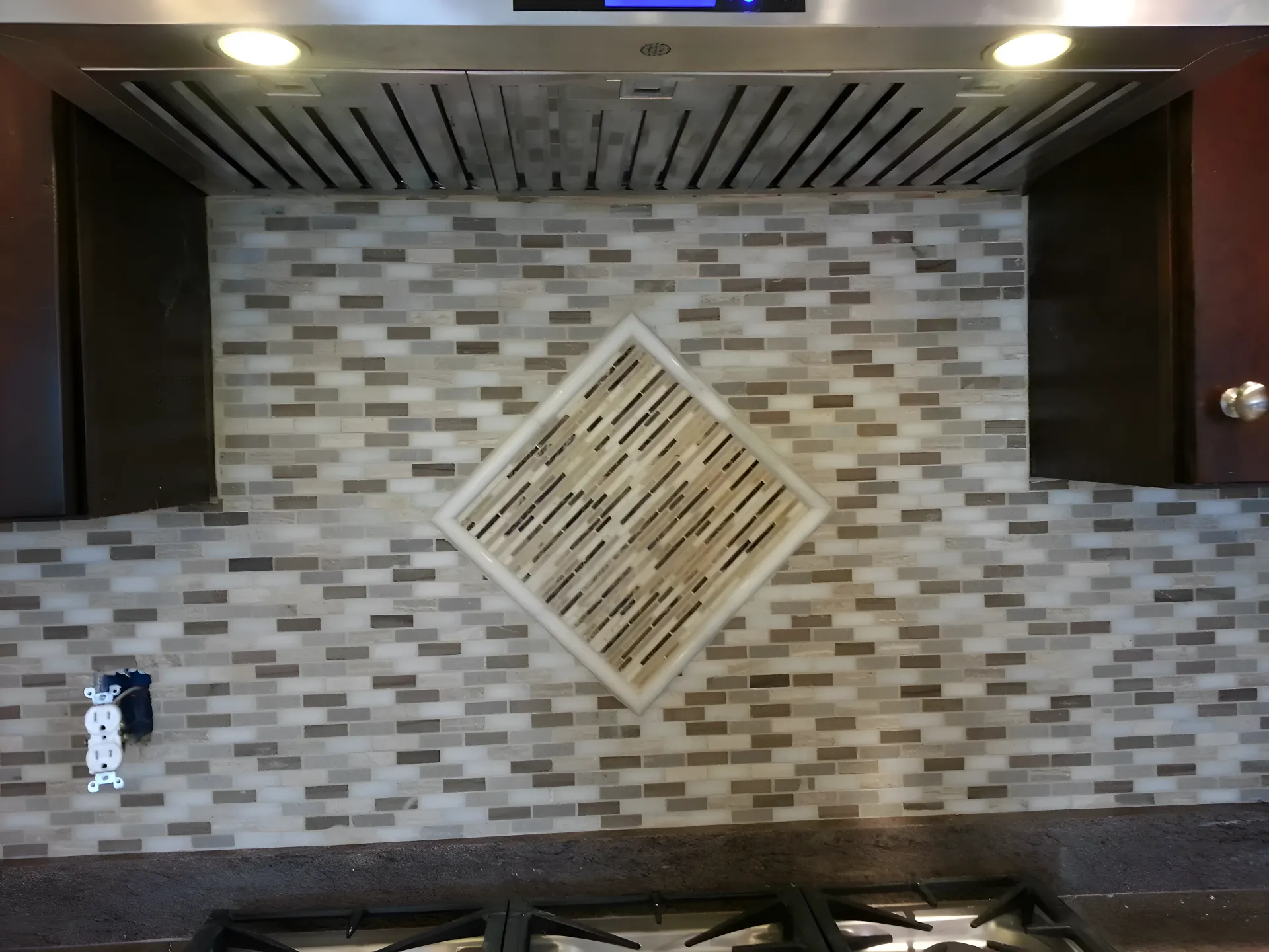 Kitchen backsplash with gray and white brick-like tile, central diamond detail, under stainless steel range hood.