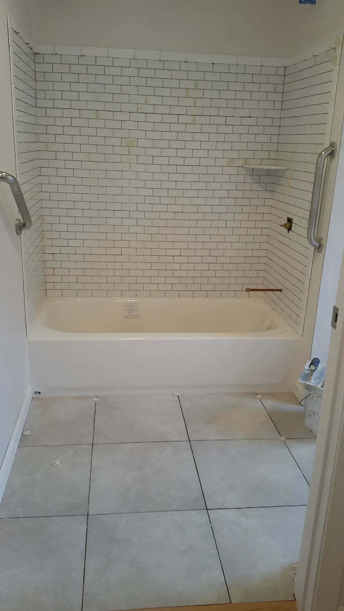 Bathroom renovation: white tiled shower surround, grab bars, and tub. Gray floor tiles are installed.