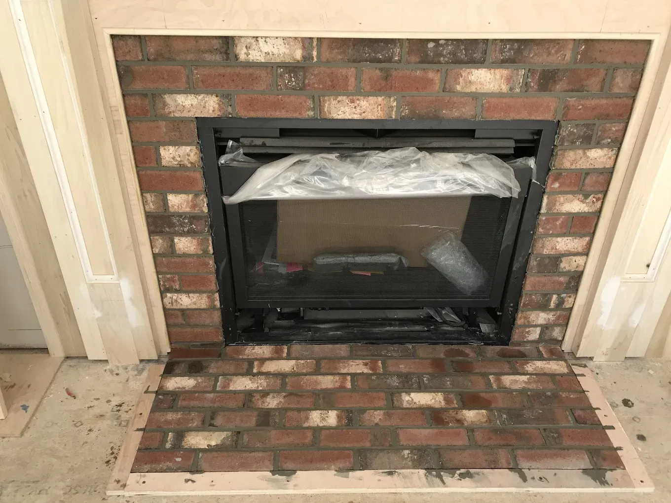 Brick fireplace with black insert, wrapped in plastic, and wooden trim.