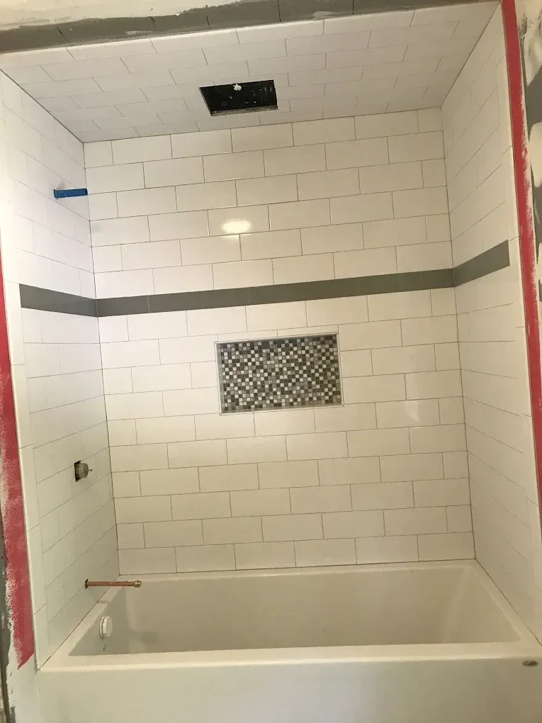 White tiled shower stall with grey accent stripe and built-in mosaic tile niche.